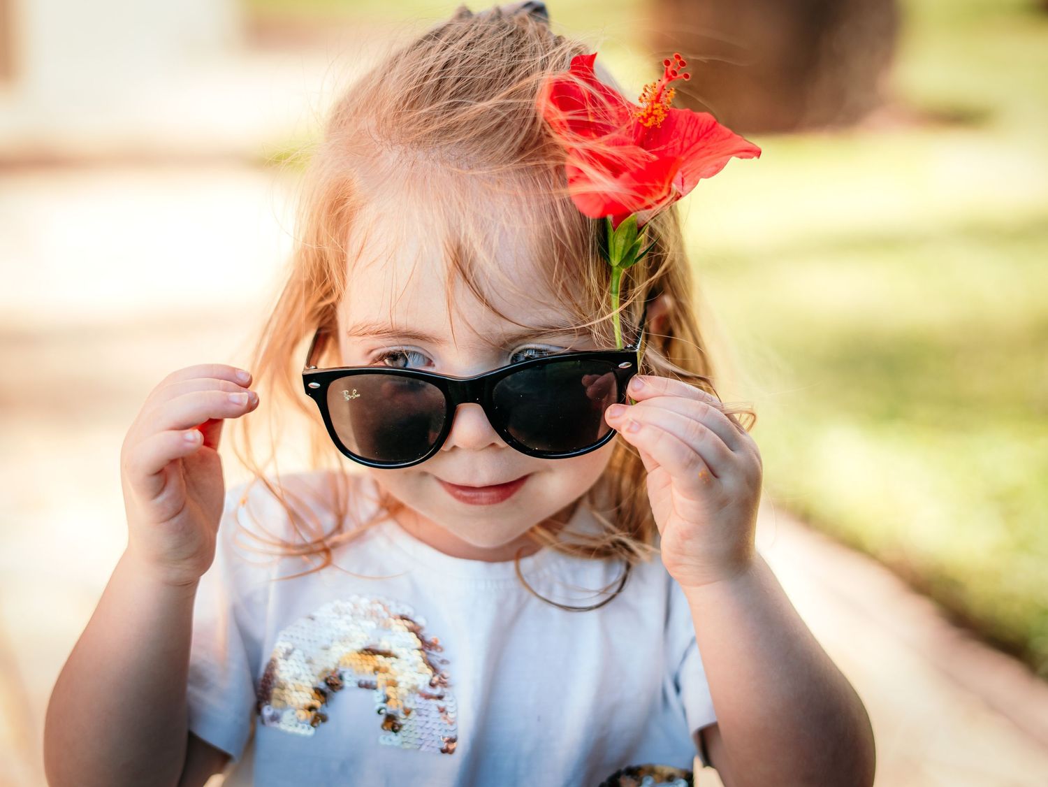 Cute little girl wearing sunglasses with a red hibiscus flower tucked behind her ear outdoors.
