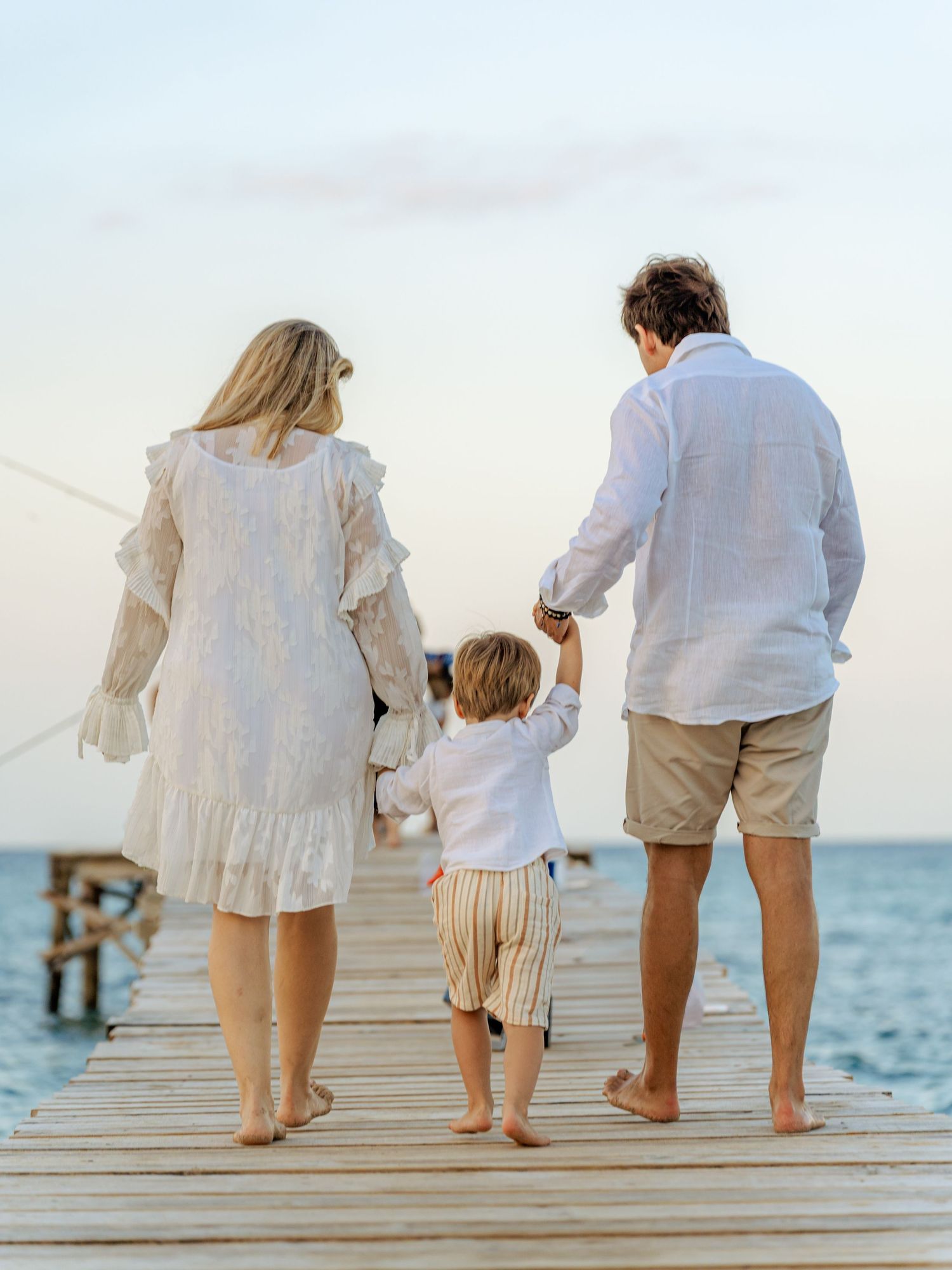 Family of three walking barefoot on wooden pier holding hands at sunset near calm blue water.