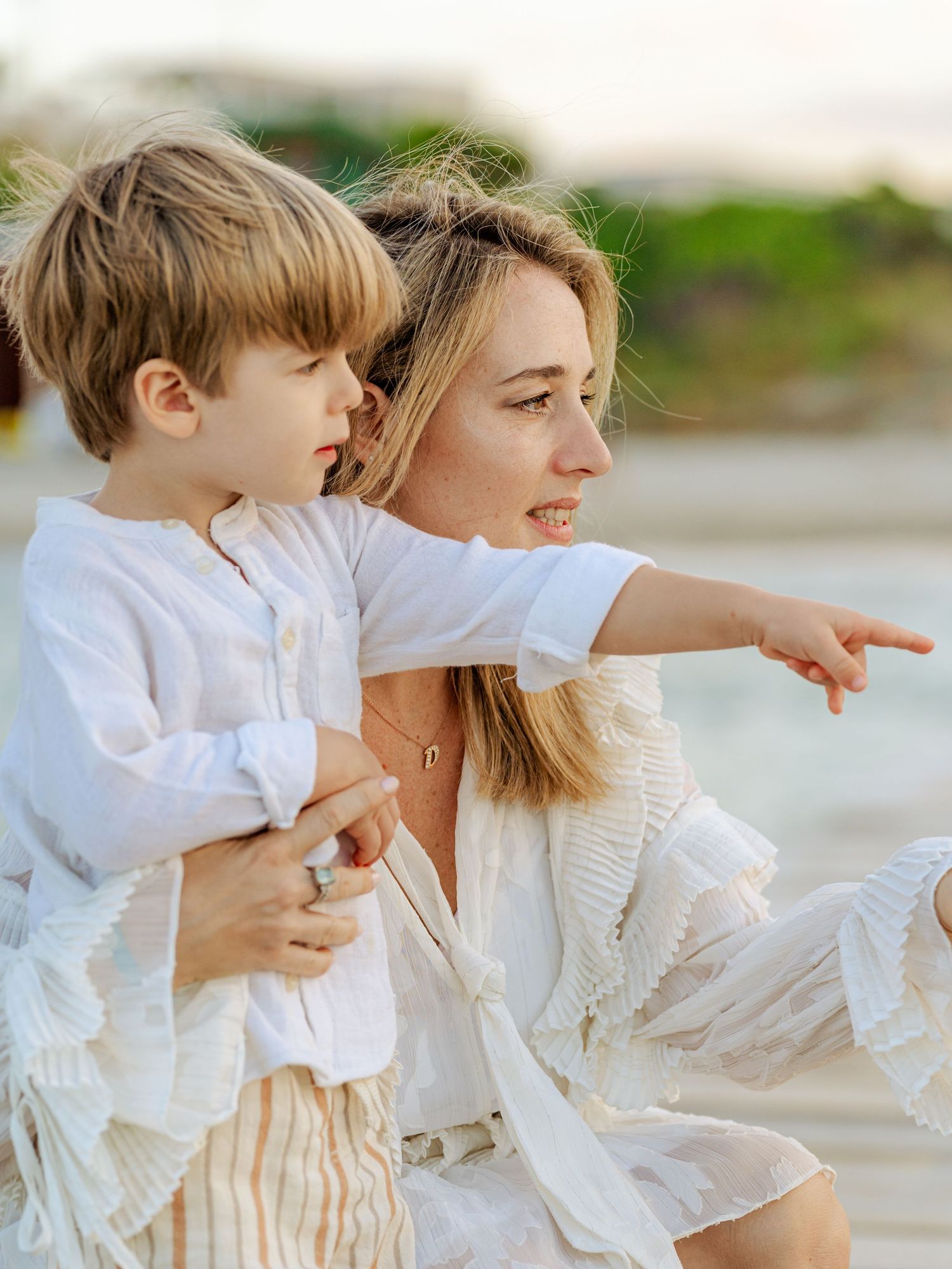 Blonde mother in white dress holding toddler son while pointing at the beach during sunset.