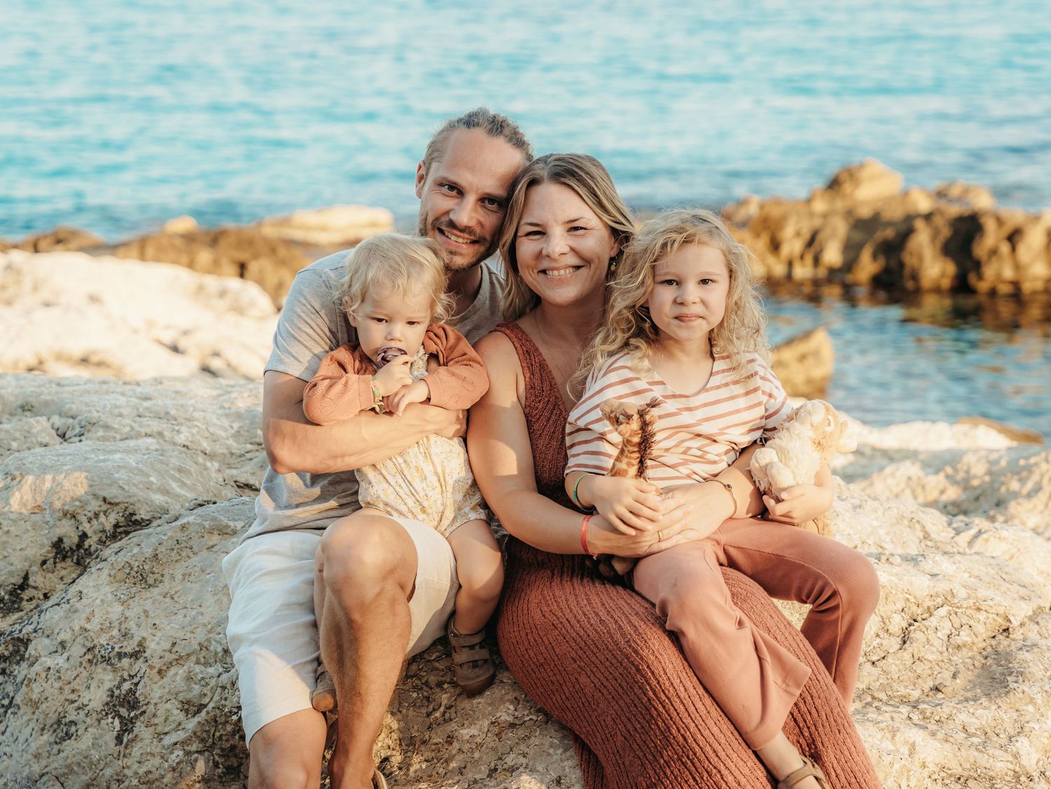 Happy family of four sitting on rocky shore by turquoise sea, parents holding toddlers in warm earth-tone outfits.
