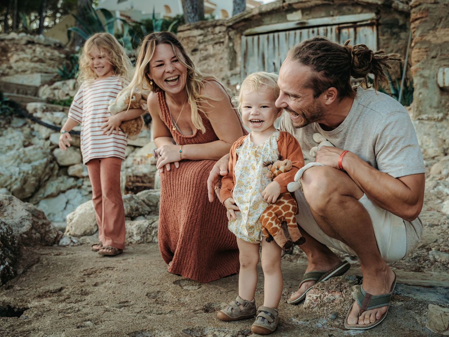 A happy family of four smiles together on sandy rocky ground with lush greenery behind them.