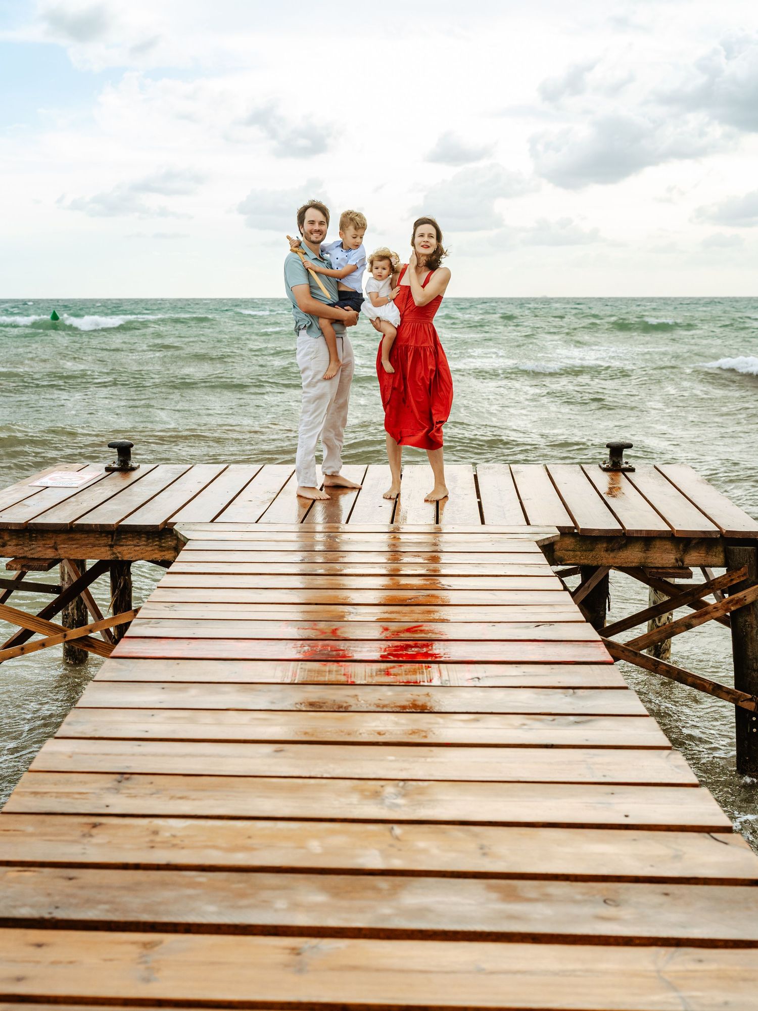 Smiling family of four on a wet wooden seaside pier, father and mother each holding a young child.