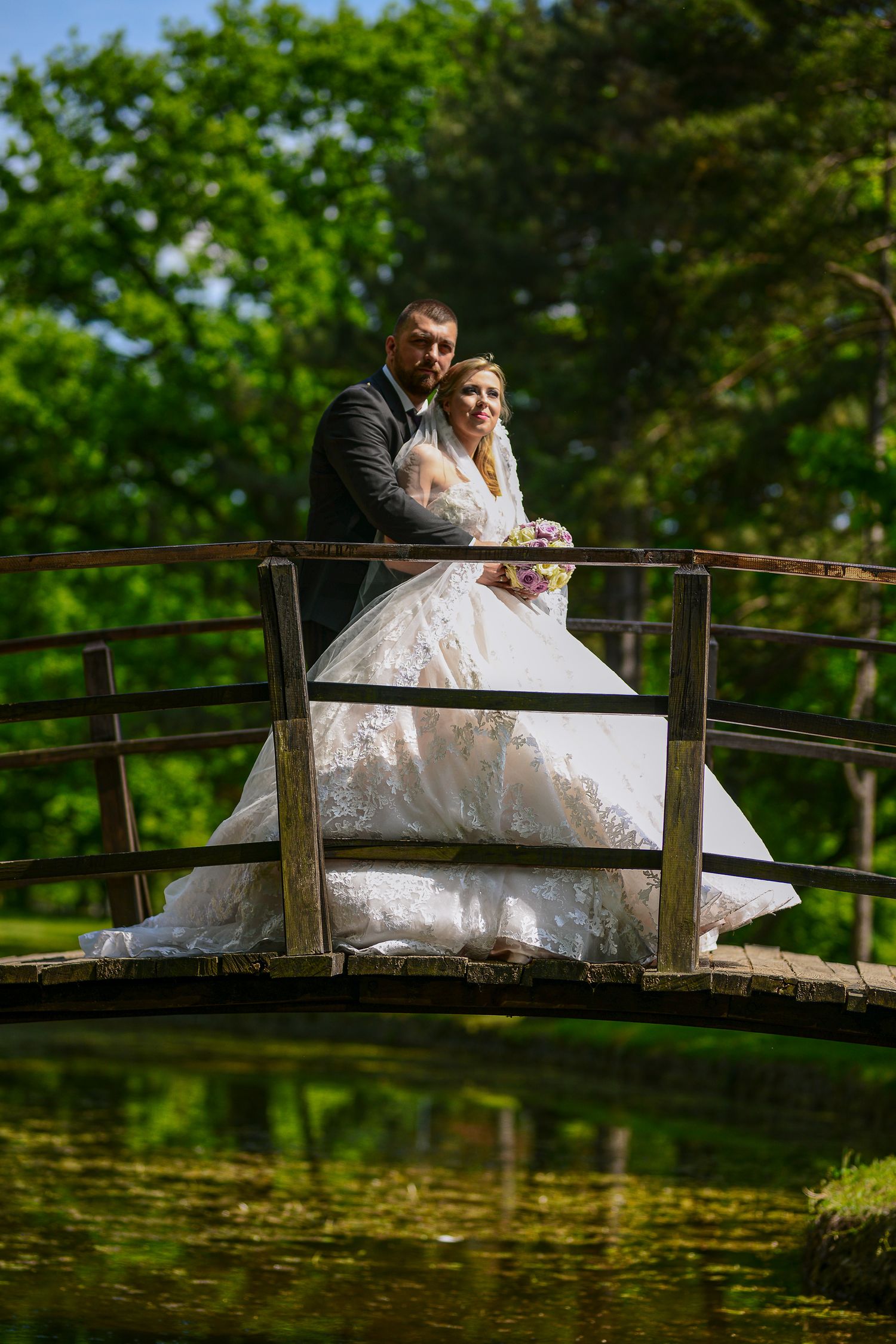 Bride and groom embrace on a wooden arched bridge over a pond surrounded by lush greenery.