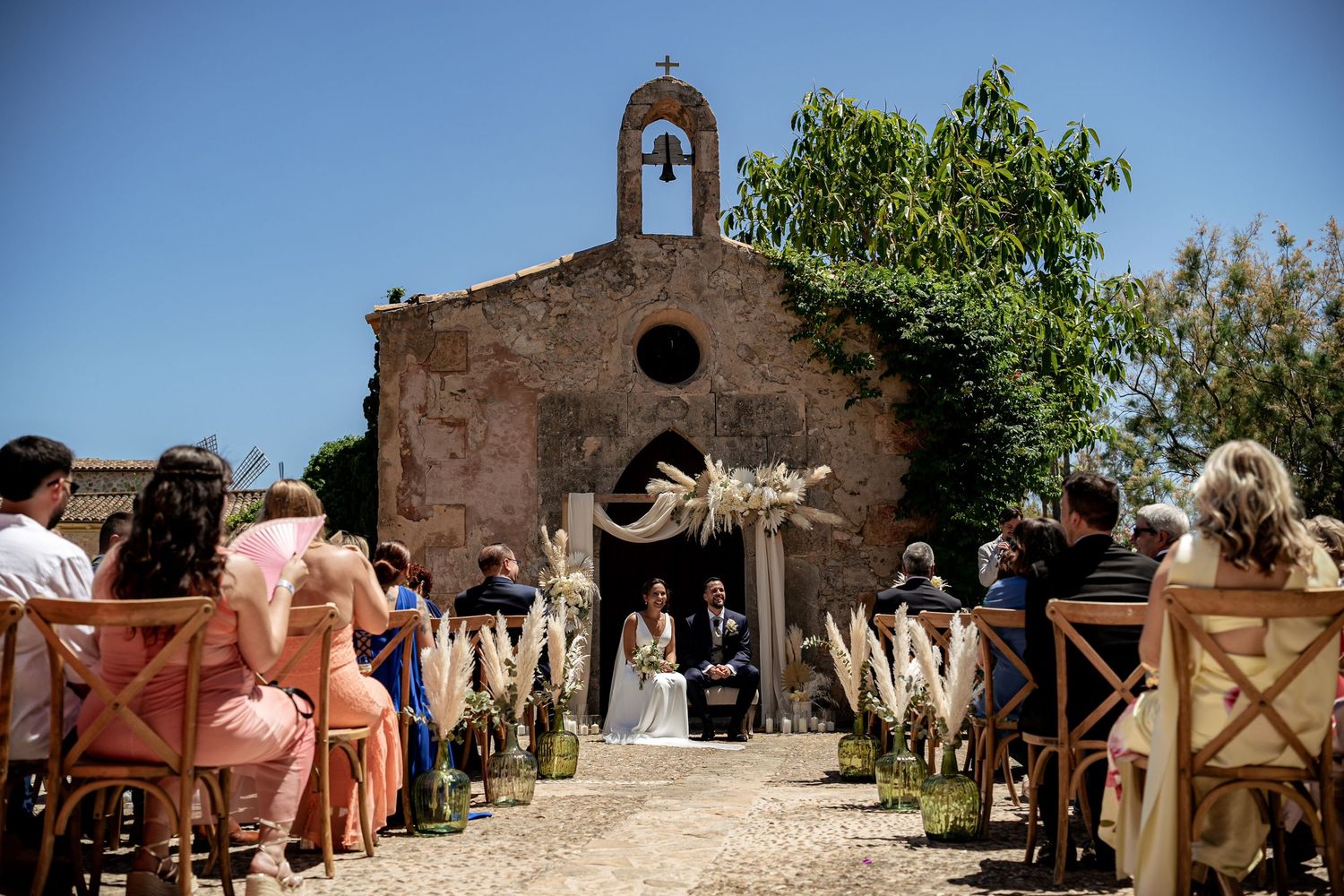 Outdoor wedding ceremony before a rustic stone chapel adorned with pampas grass floral arrangements.