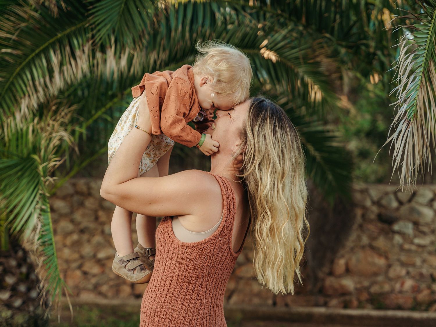 Blonde mother in rust dress lifting and kissing her baby near tropical palm trees.