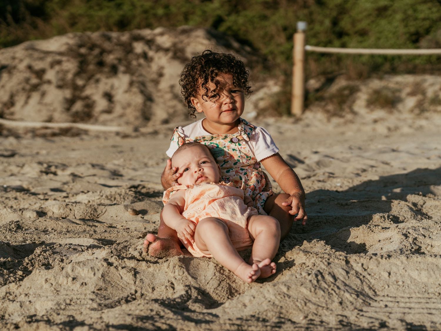 Toddler with curly hair sitting on sand gently holding a newborn baby sibling.