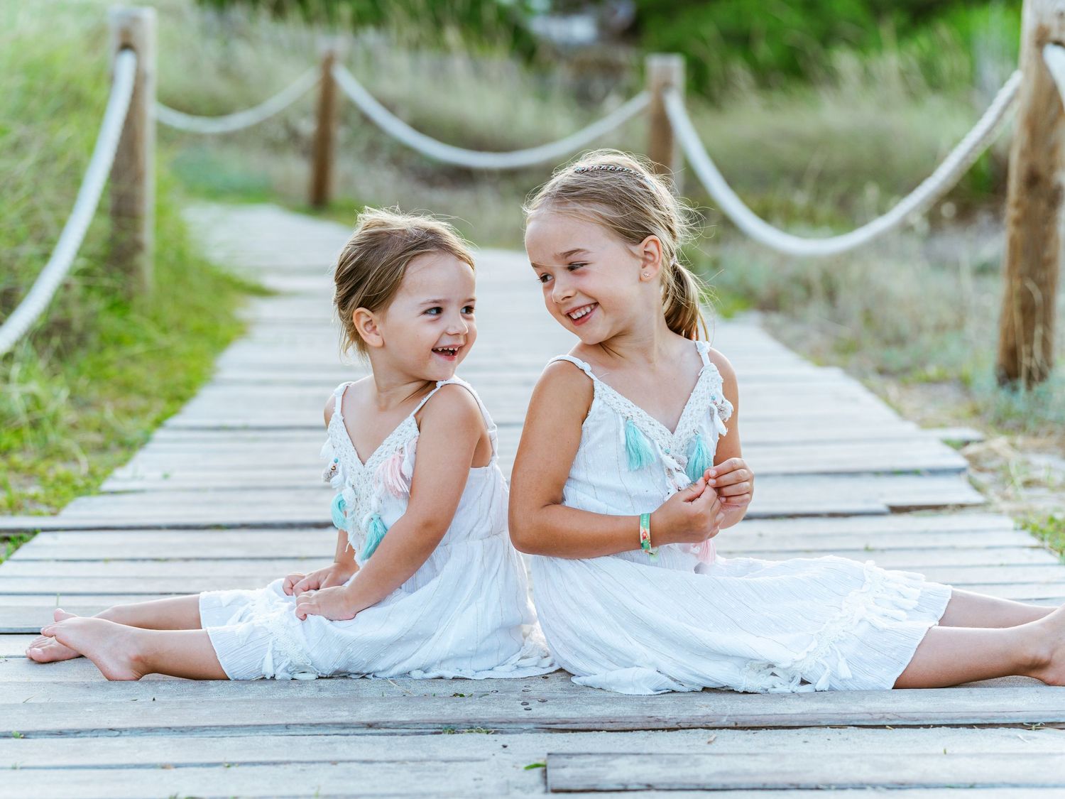 Two young girls in matching white dresses sitting back-to-back on a wooden boardwalk, smiling joyfully.