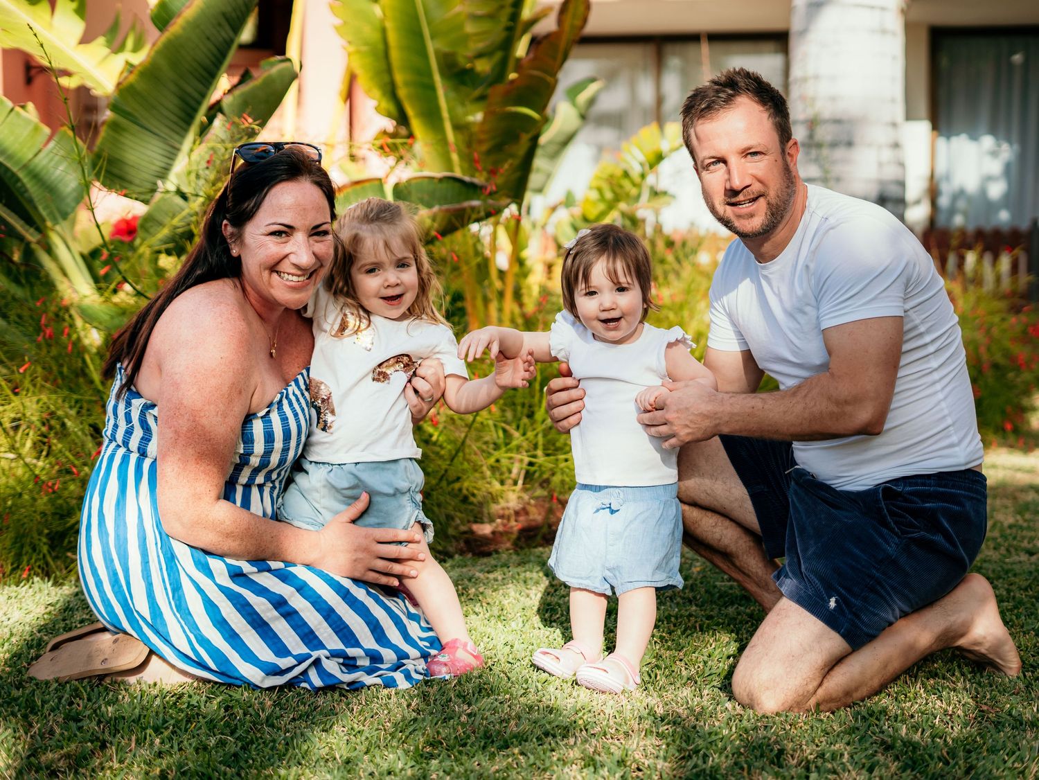 Happy family with two toddler girls posing together on green grass in tropical garden.