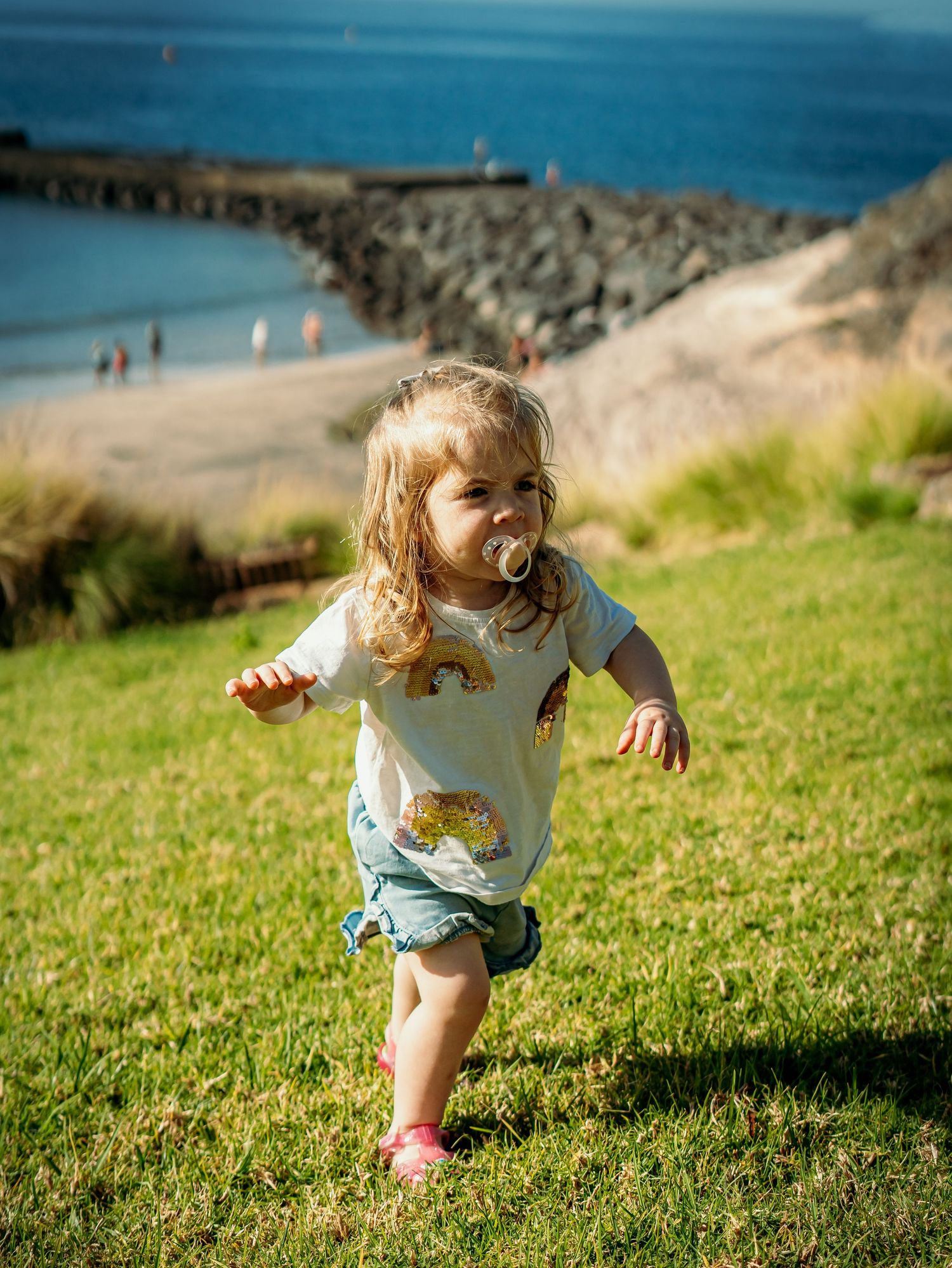 Energetic toddler girl with pacifier running on green hillside with ocean and harbor behind her.