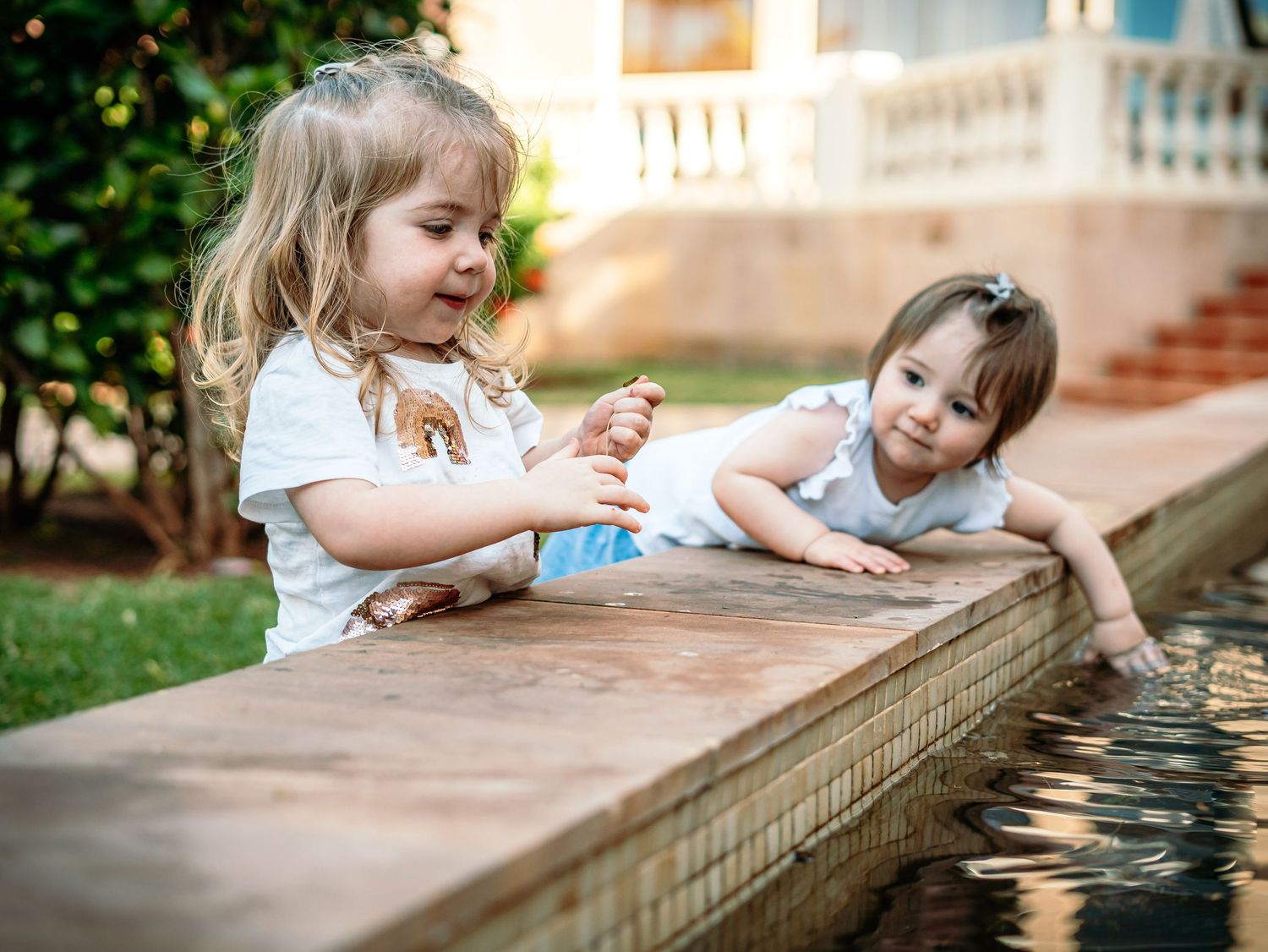 Two young sisters joyfully playing together at a garden water feature on sunny day.