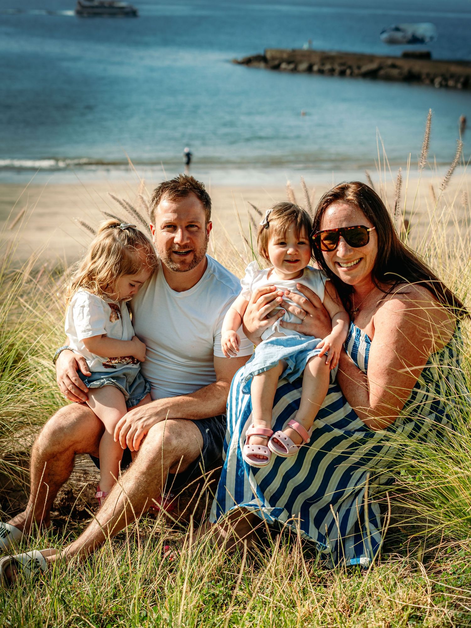 A happy family of four with two toddlers poses together on grassy coastal hillside by the sea.