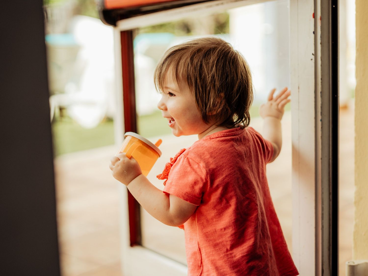 Happy toddler girl in red shirt holding cup while looking out an open glass door outside.