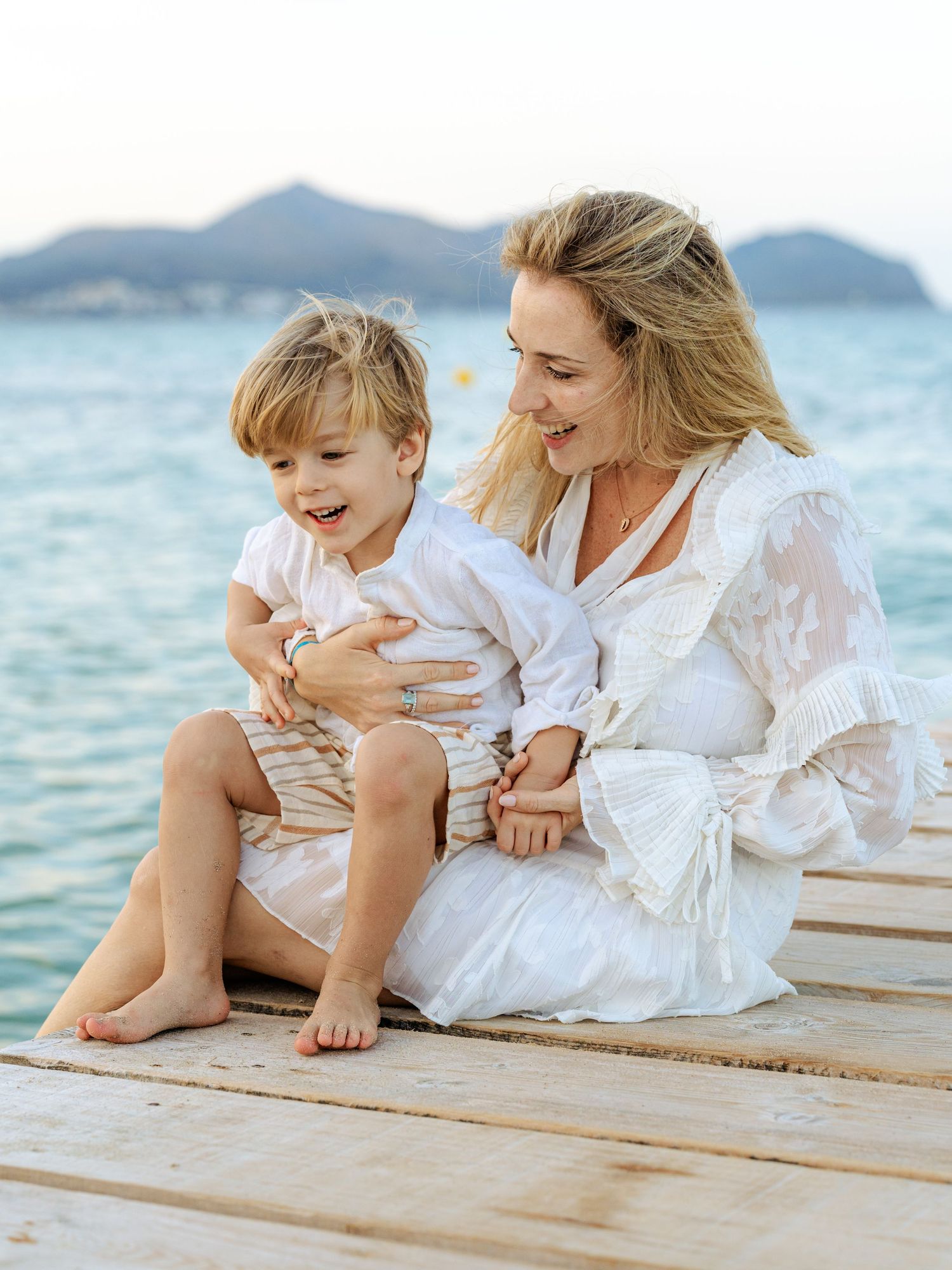 Joyful mother in white dress laughing with her happy toddler son on a seaside wooden pier.