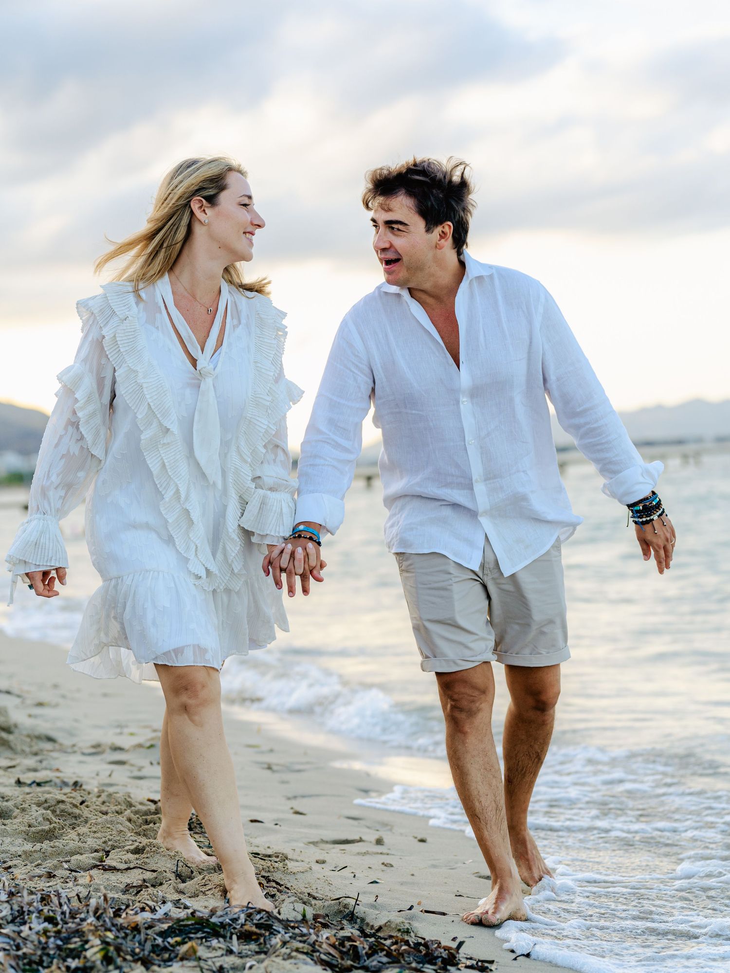 Smiling couple in white outfits laughing together while walking barefoot on a scenic beach.