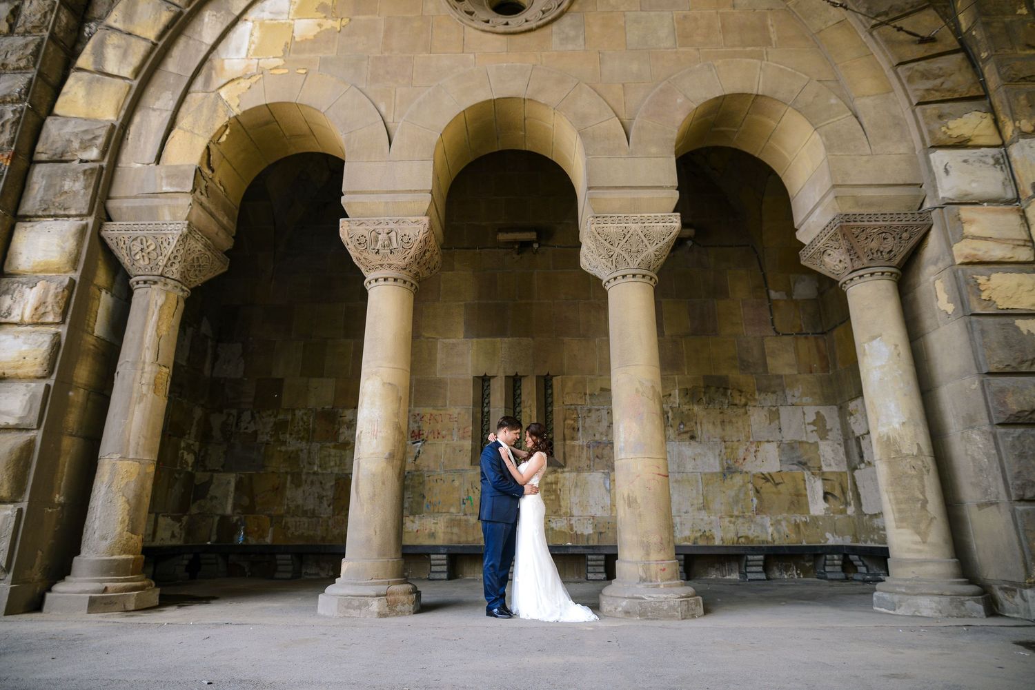 Bride and groom kissing beneath grand stone arches of historic building in elegant wedding portrait.