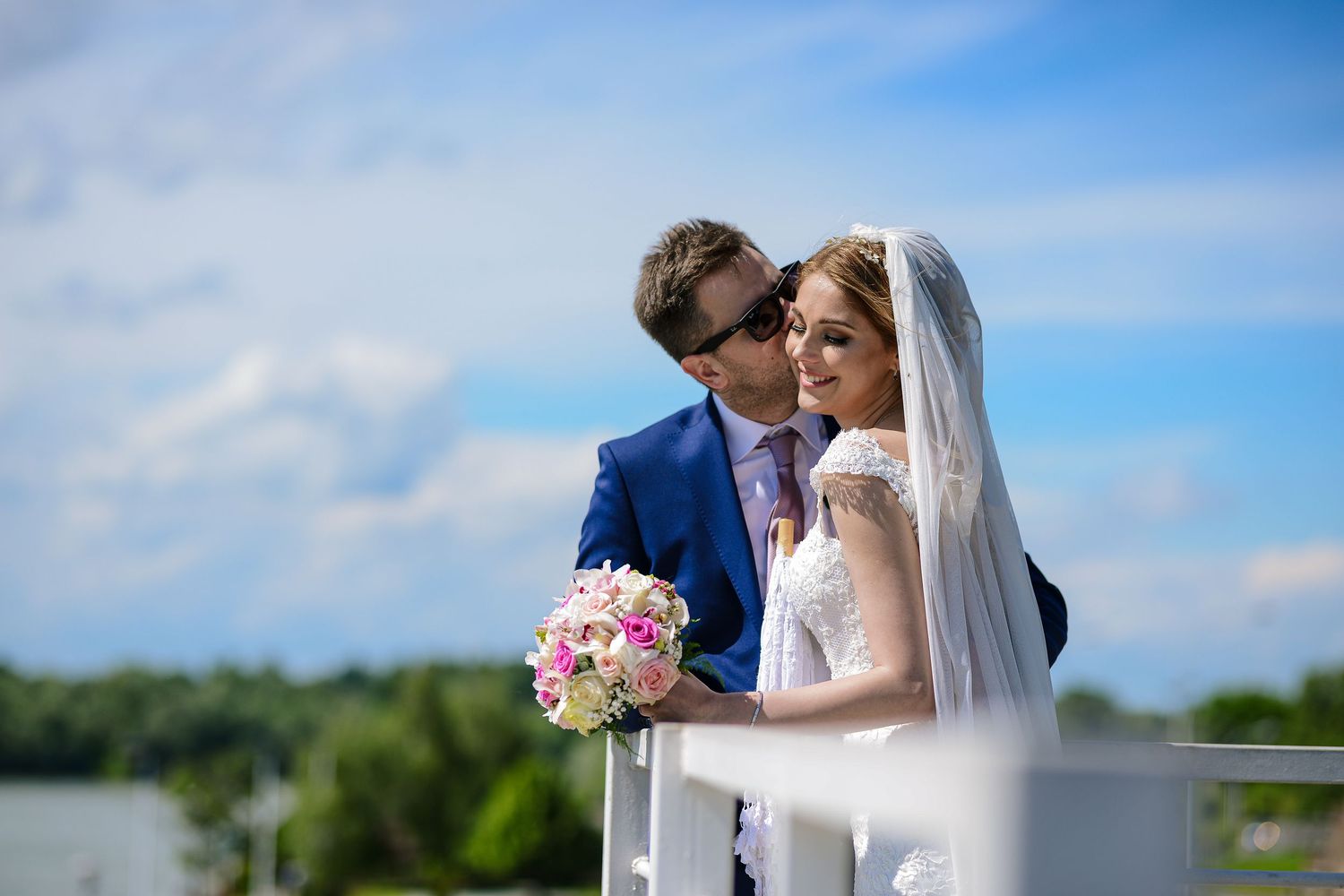 Newlyweds in blue suit and white gown with veil pose on a balcony under a bright blue sky.
