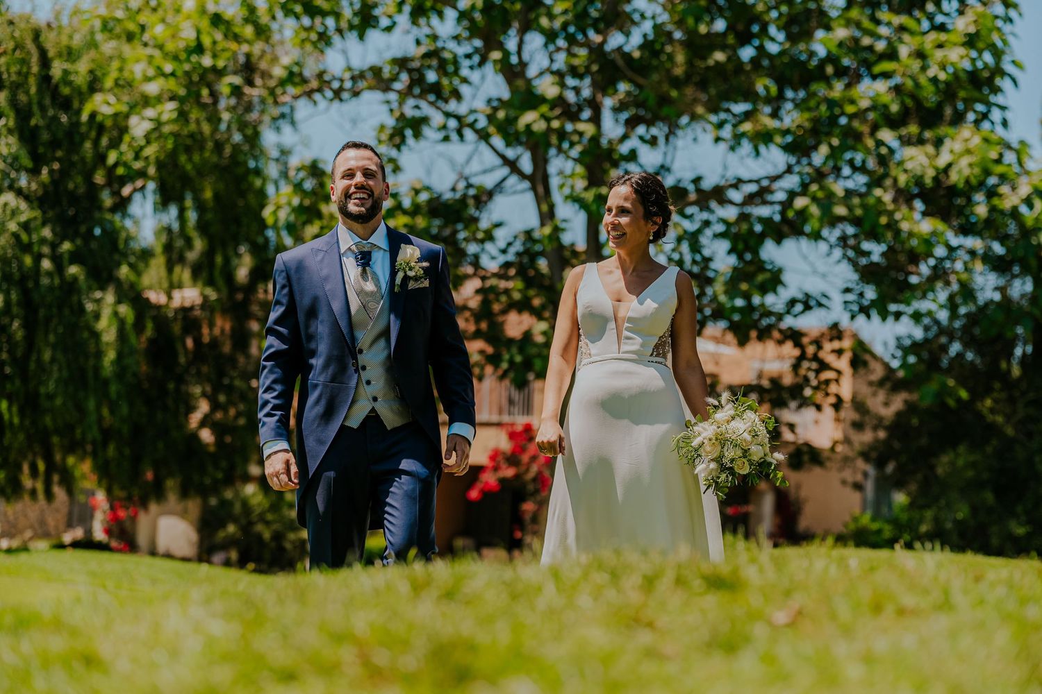 Smiling bride and groom walking hand in hand through lush green garden on their wedding day.