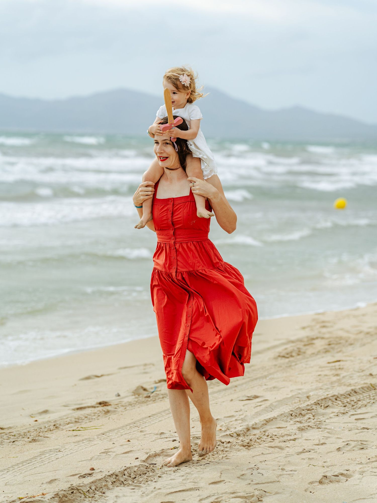 Woman in red dress carries toddler on shoulders while walking barefoot on sandy beach.