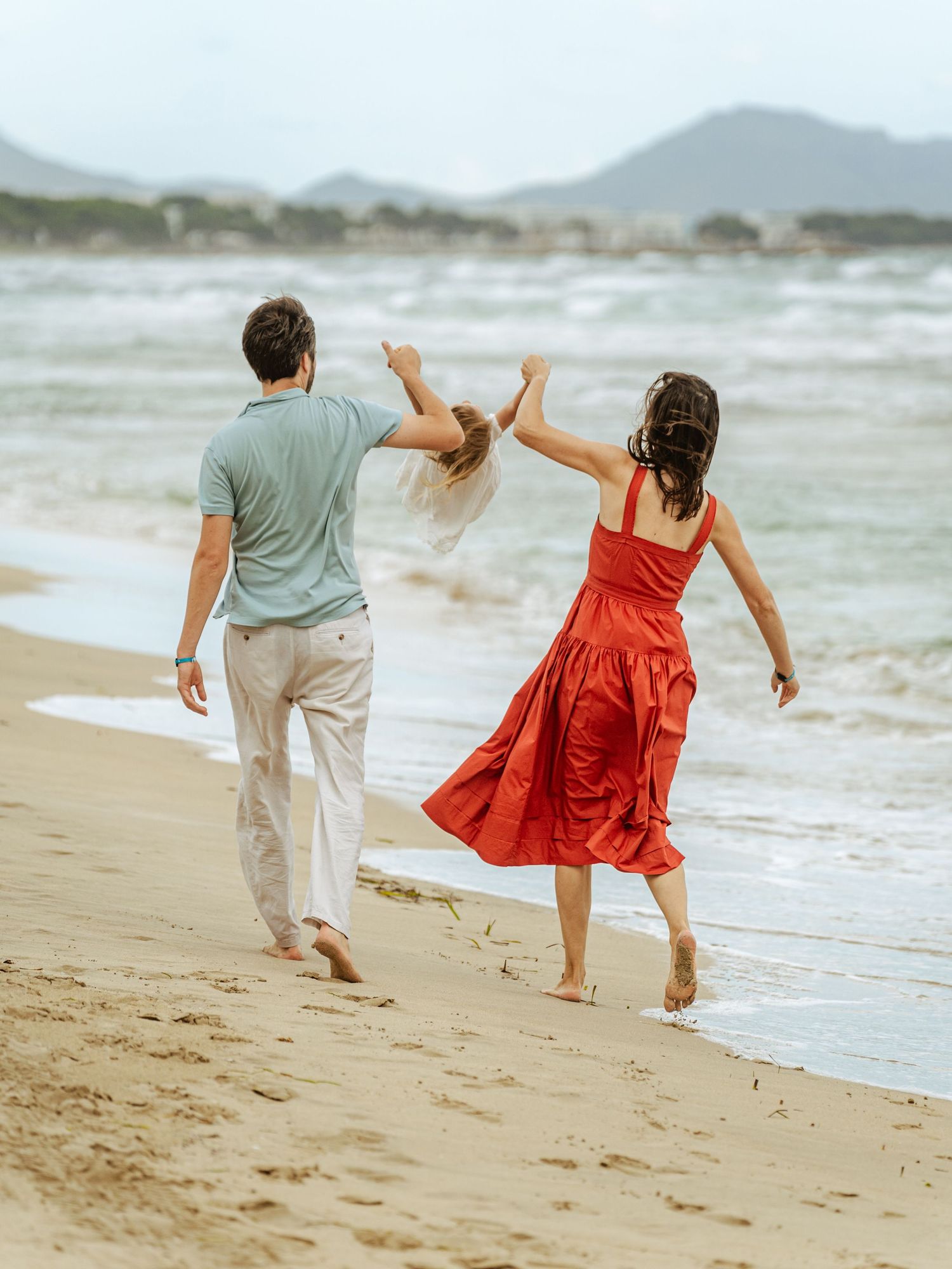 Family walking on beach, parents swinging child between them near ocean waves.