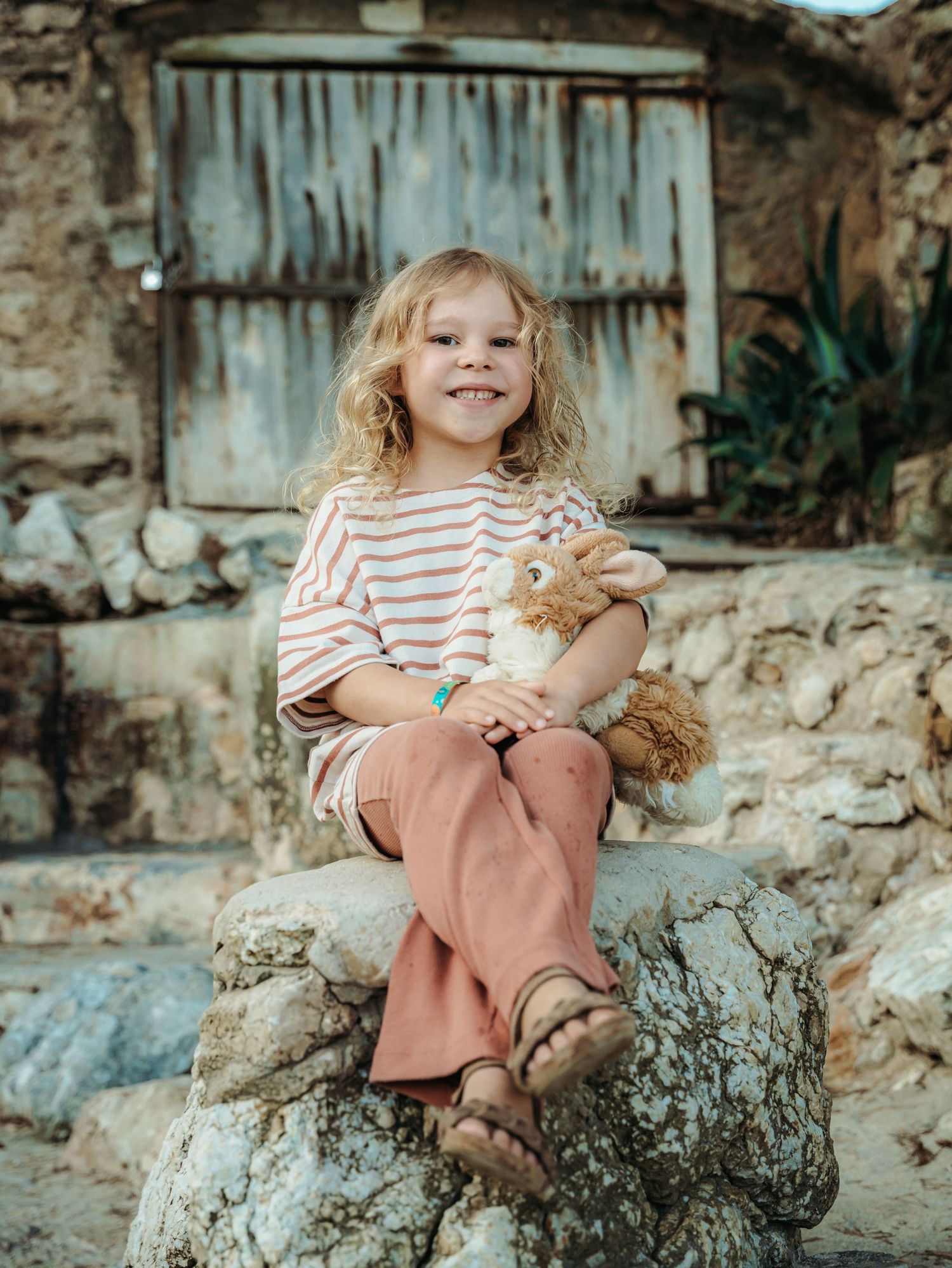Happy blonde curly-haired girl in striped top and pink pants sitting on rock holding stuffed animal toy.