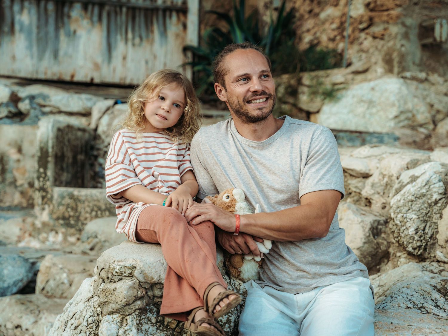 A smiling man sits on rocks holding a duckling with a young blonde child in striped shirt and rust pants.