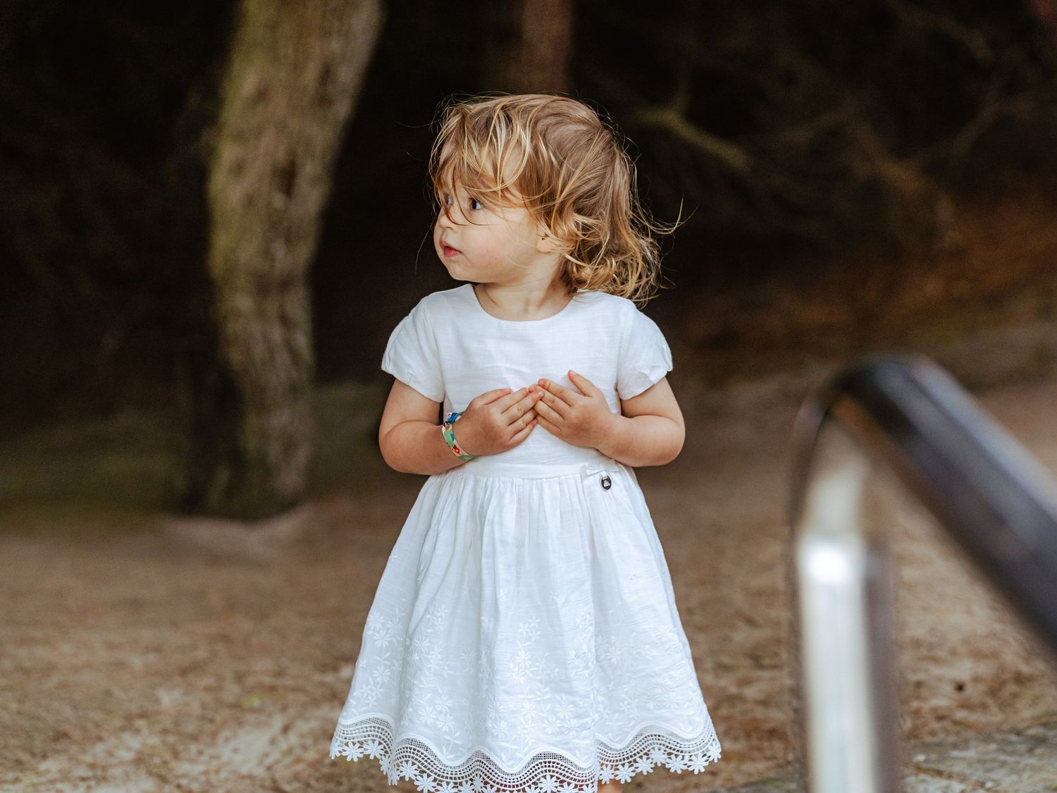 Curly-haired toddler girl in white embroidered dress standing barefoot near large tree.