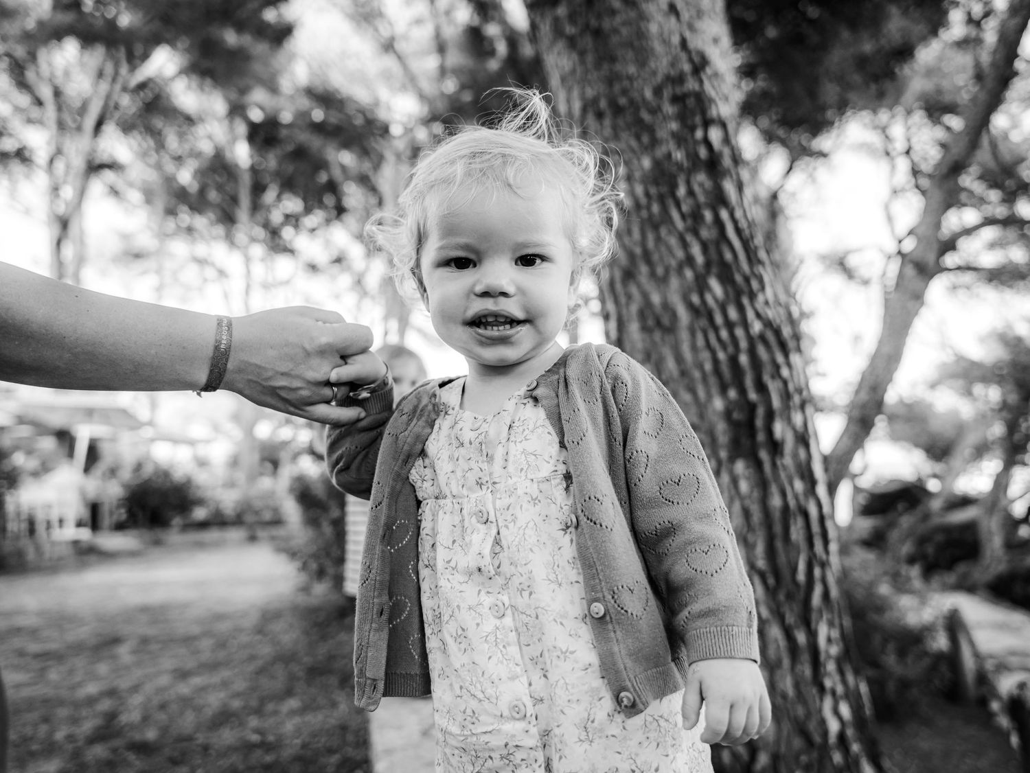 Black and white photo of smiling toddler girl holding adult hand near pine trees outdoors.