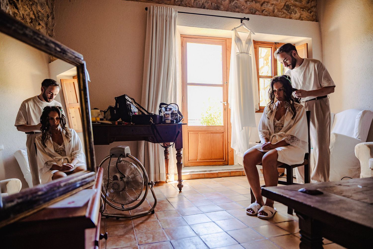 Bride and bridesmaids getting ready in rustic room with hair styling and mirror reflections.