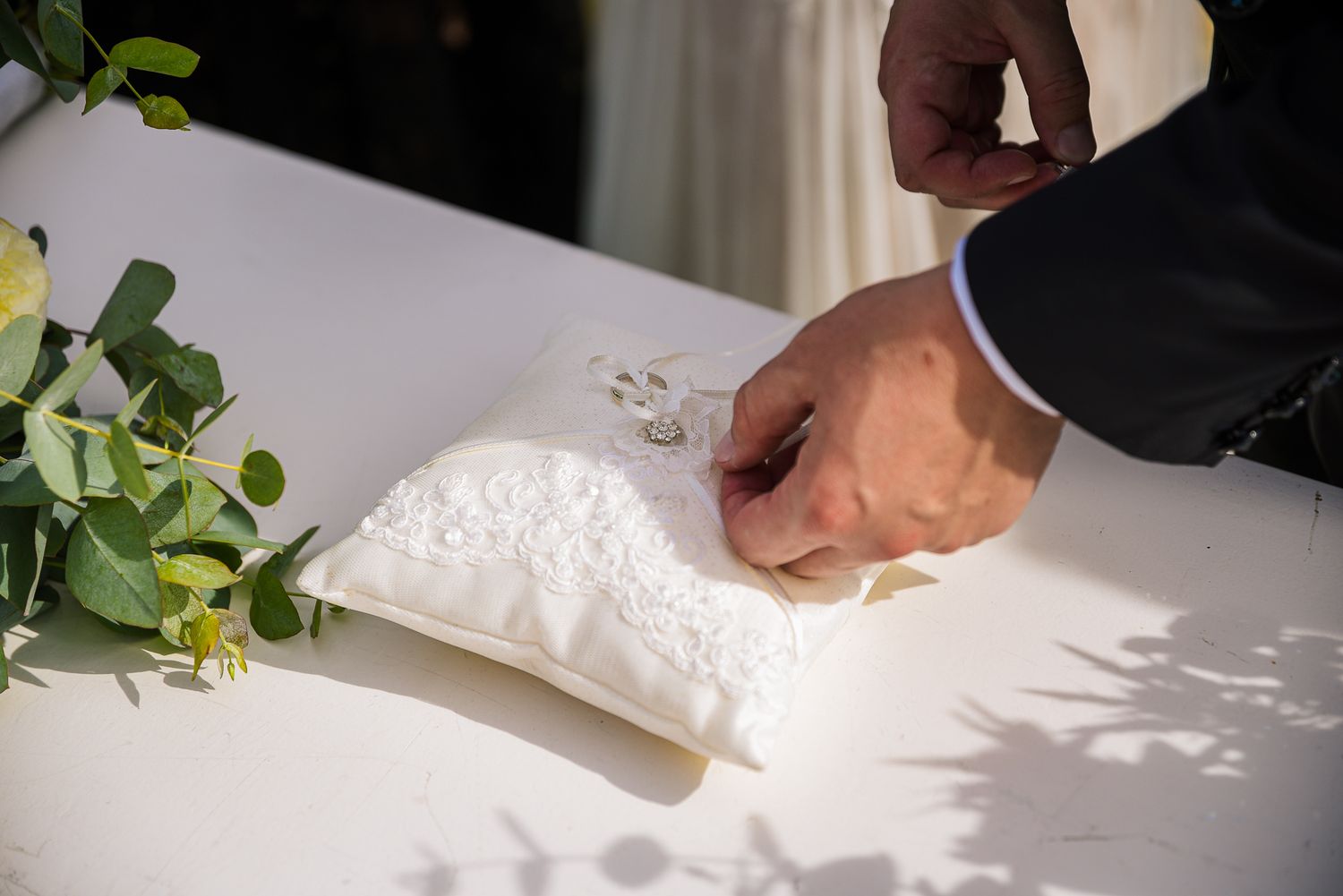 Groom picks up wedding rings from white lace pillow on table during ceremony.