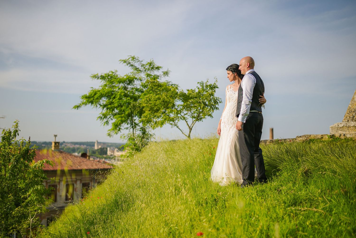 Bride and groom stand together on a green hillside with a scenic town and blue sky backdrop.