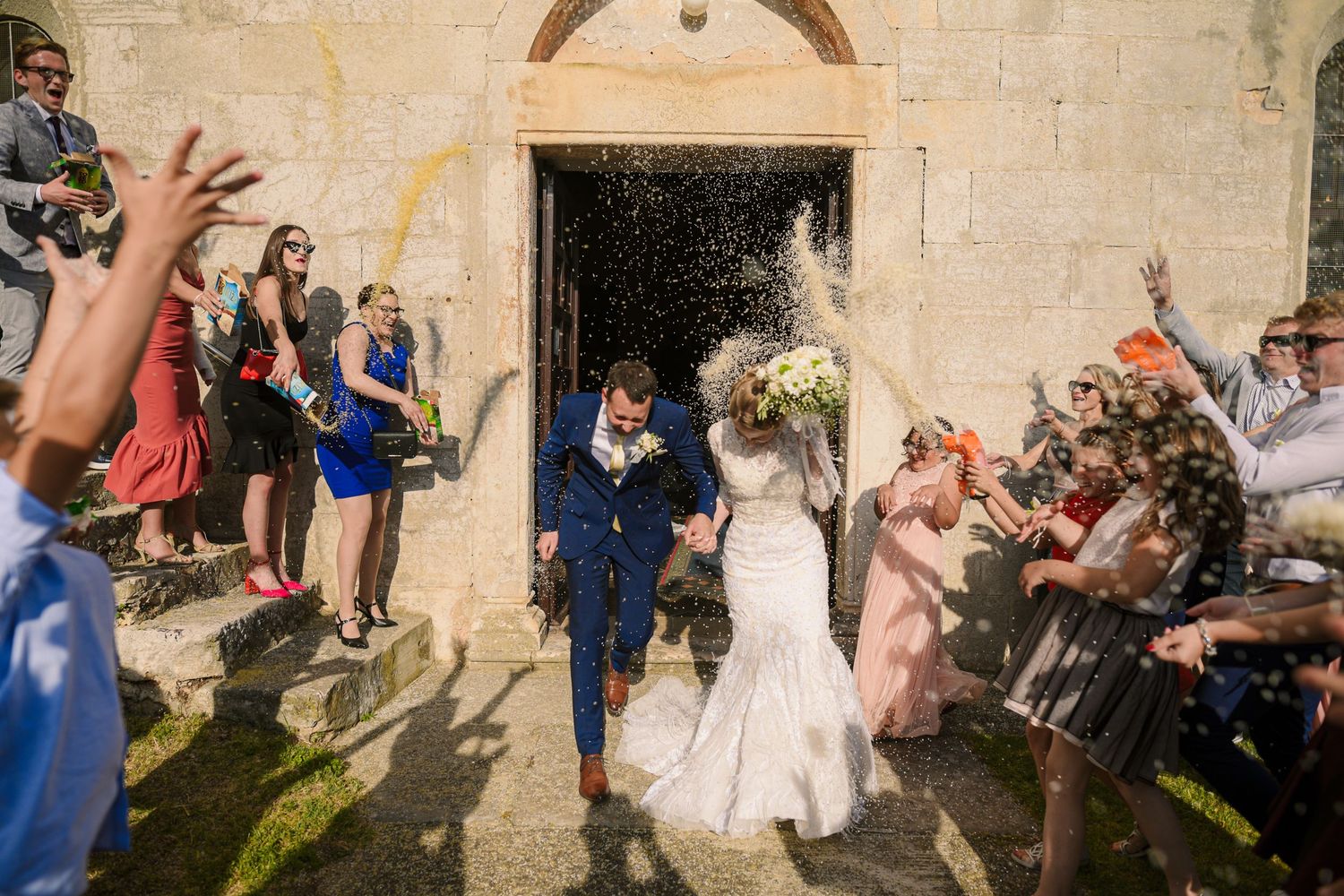 Newlyweds exit stone church as guests celebrate with confetti shower in bright sunlight.