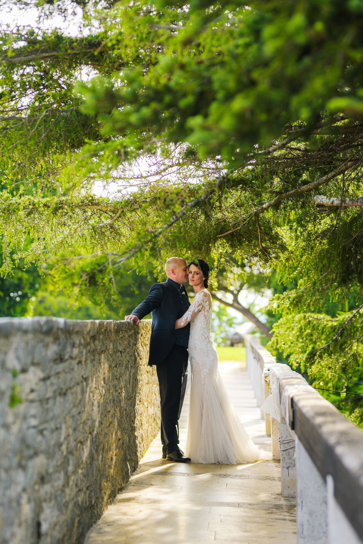 Bride in white lace gown and groom in dark suit pose on a sunlit stone bridge walkway.