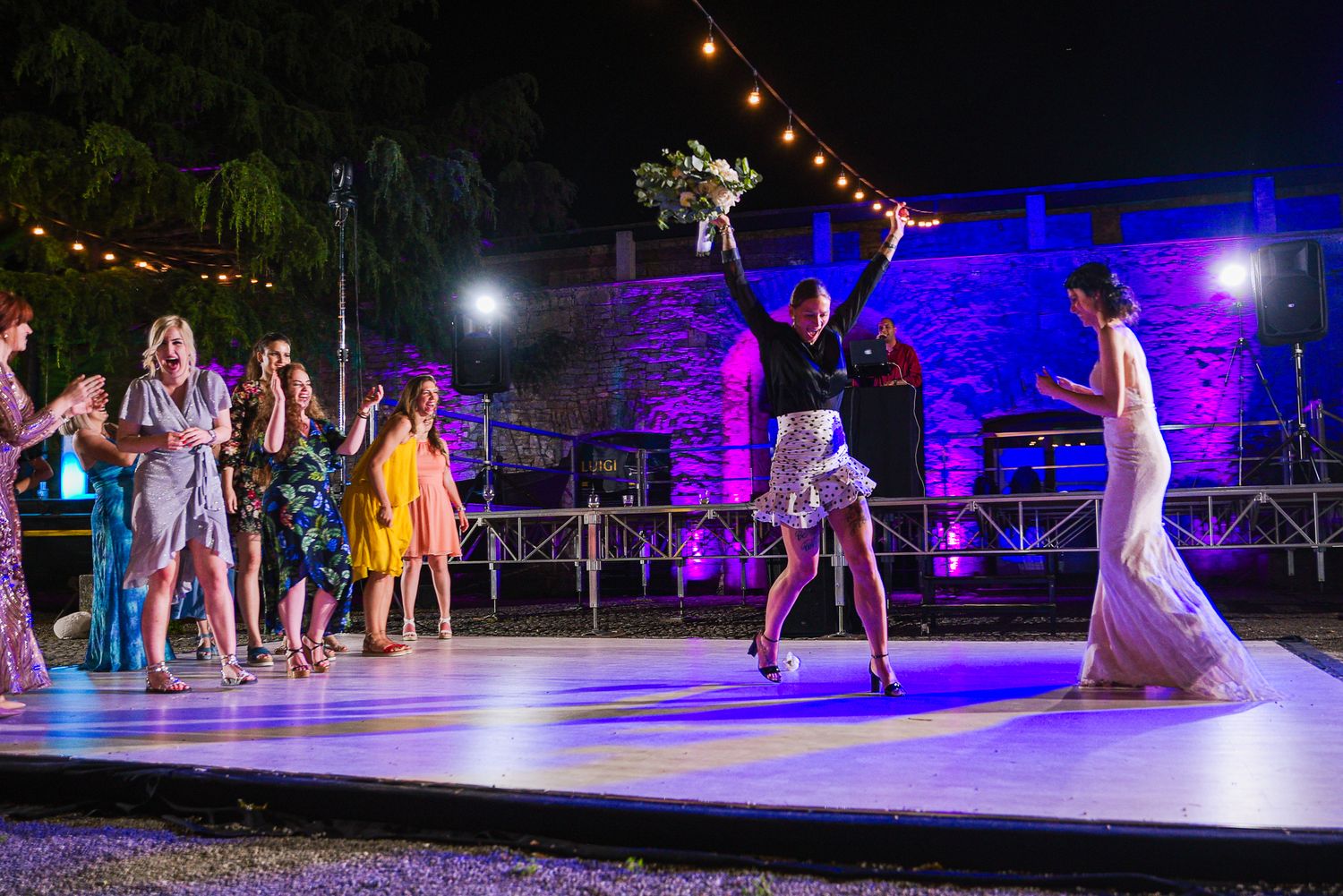 Bride tossing wedding bouquet to excited guests on outdoor stage at night.