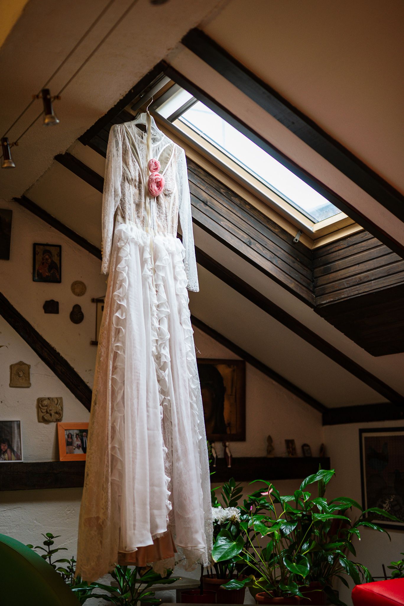 Elegant white lace wedding dress with pink floral detail hanging in a rustic attic with skylight.