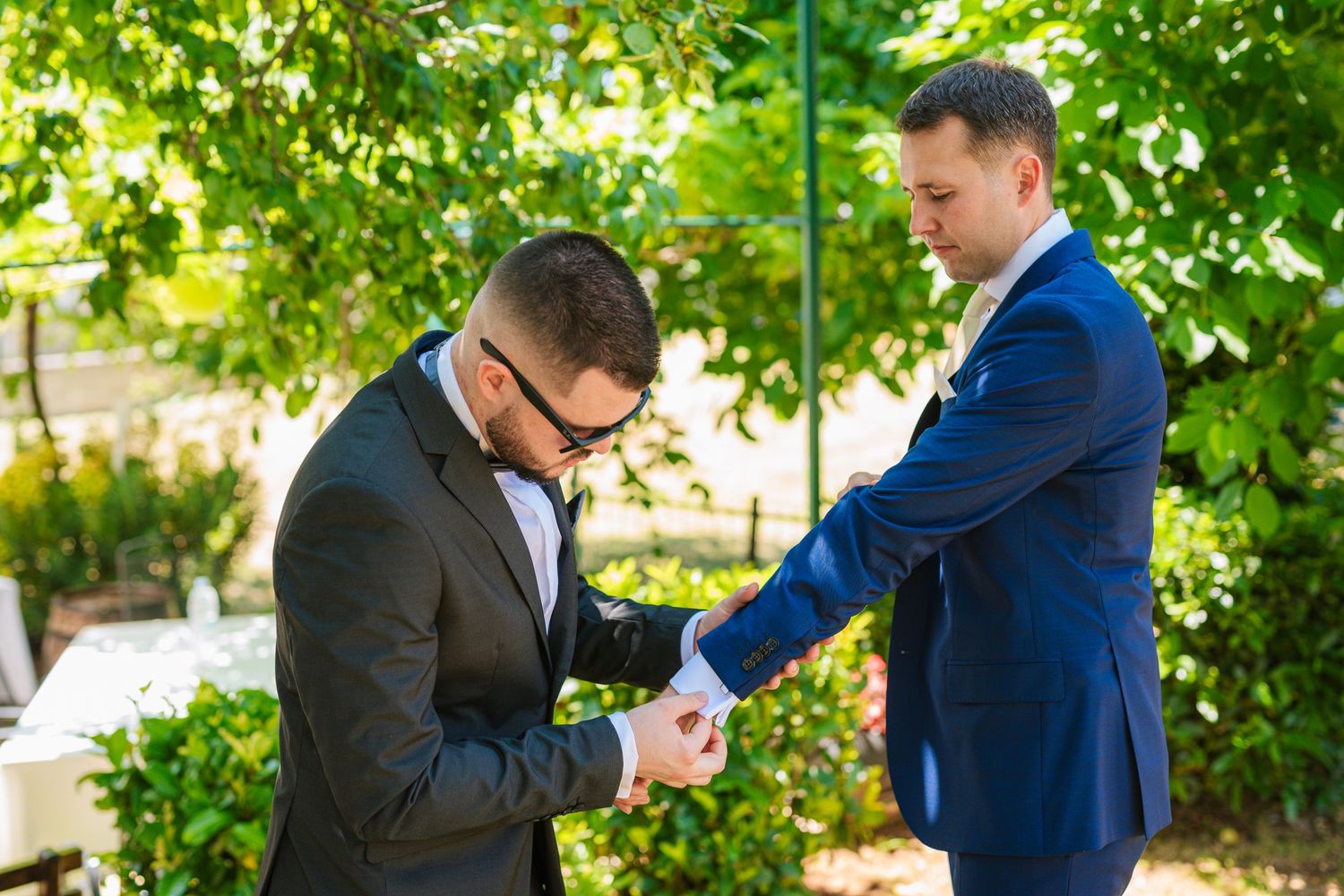 Groomsman in blue suit helps another man with cufflinks under green trees outdoors.