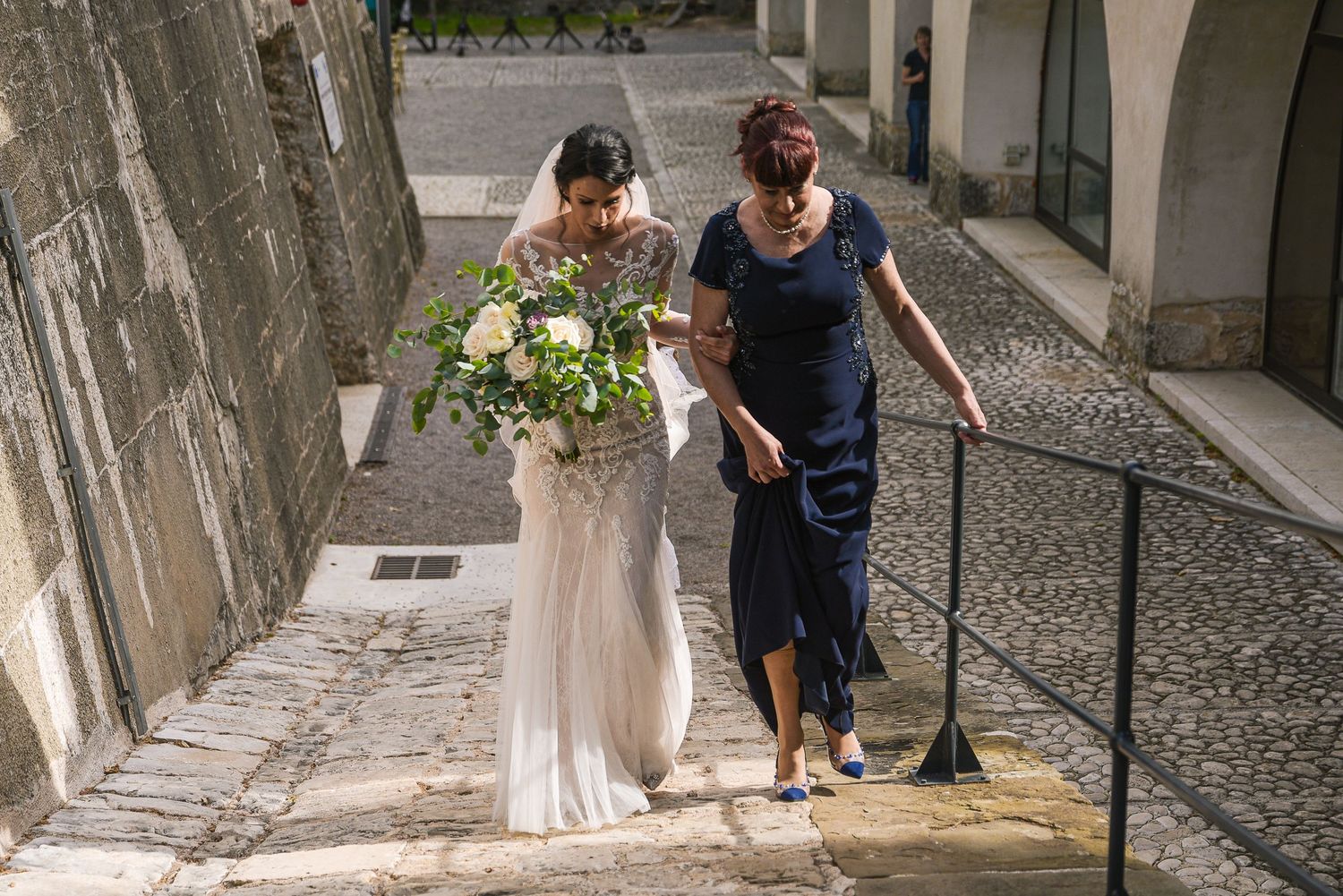 Bride in lace gown carrying bouquet ascending stone steps with female companion.