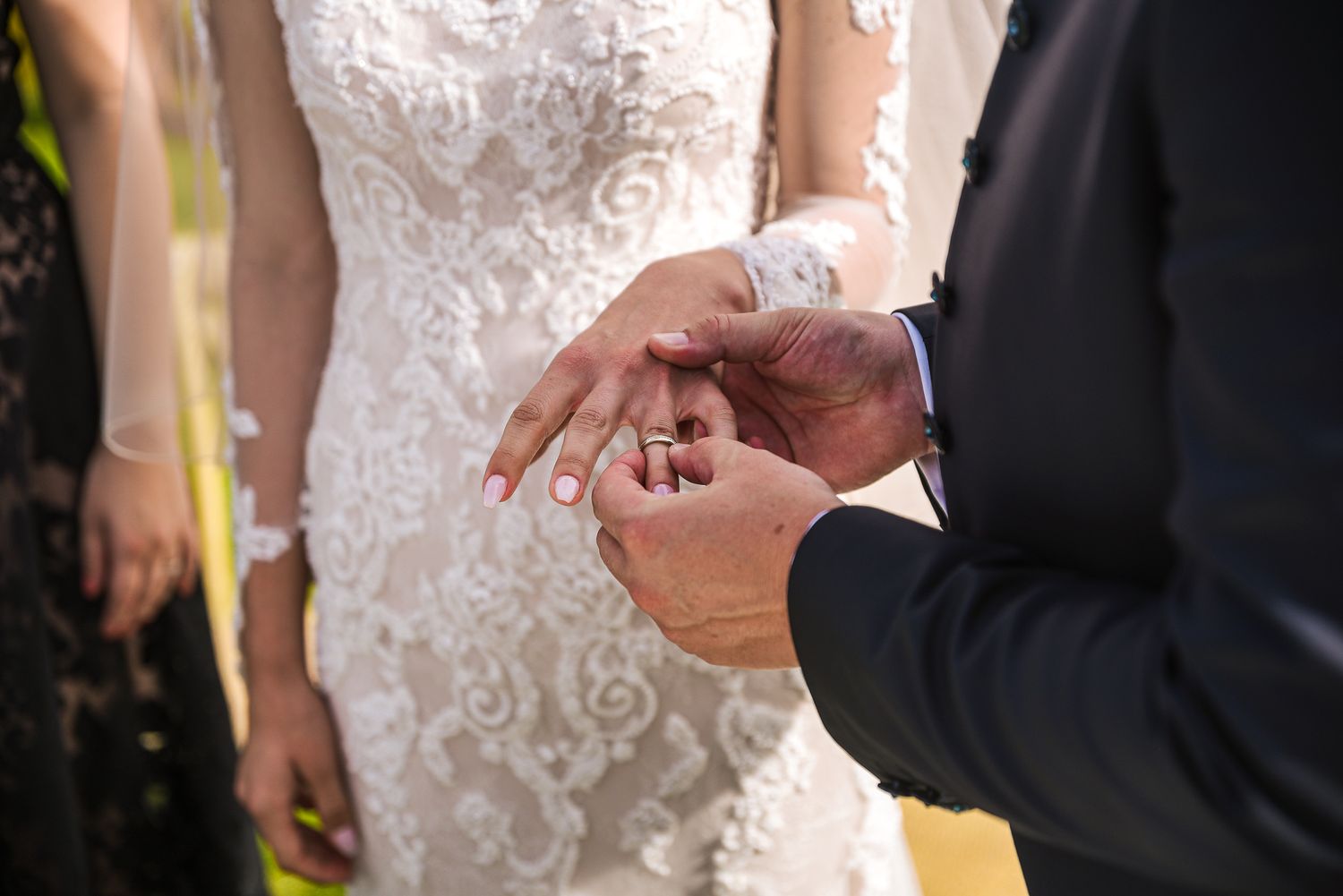 Close-up of groom sliding wedding ring onto brides finger, bride in beautiful white lace wedding gown.