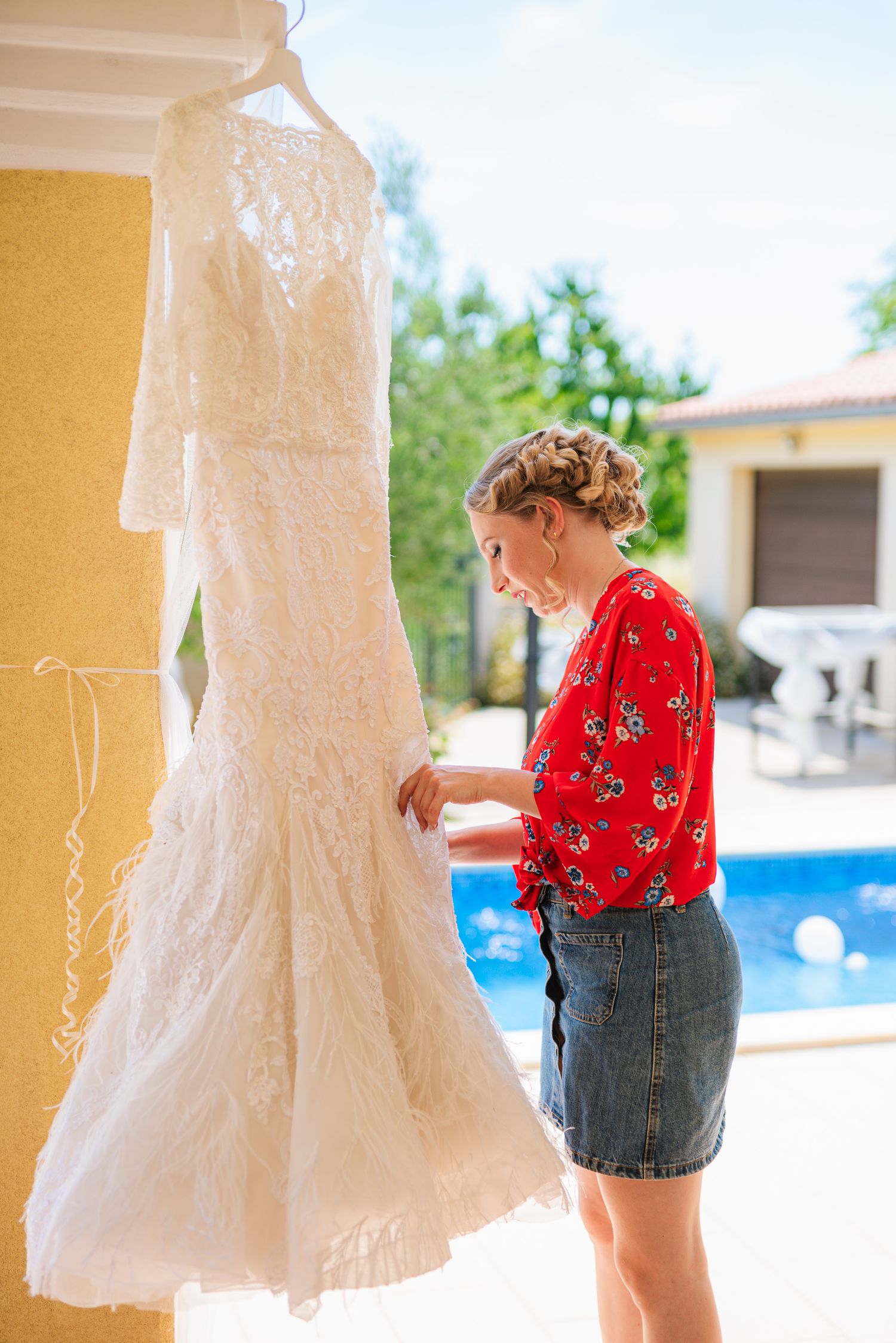Blonde bridesmaid in red top examines lace wedding dress hanging by a sunny poolside.