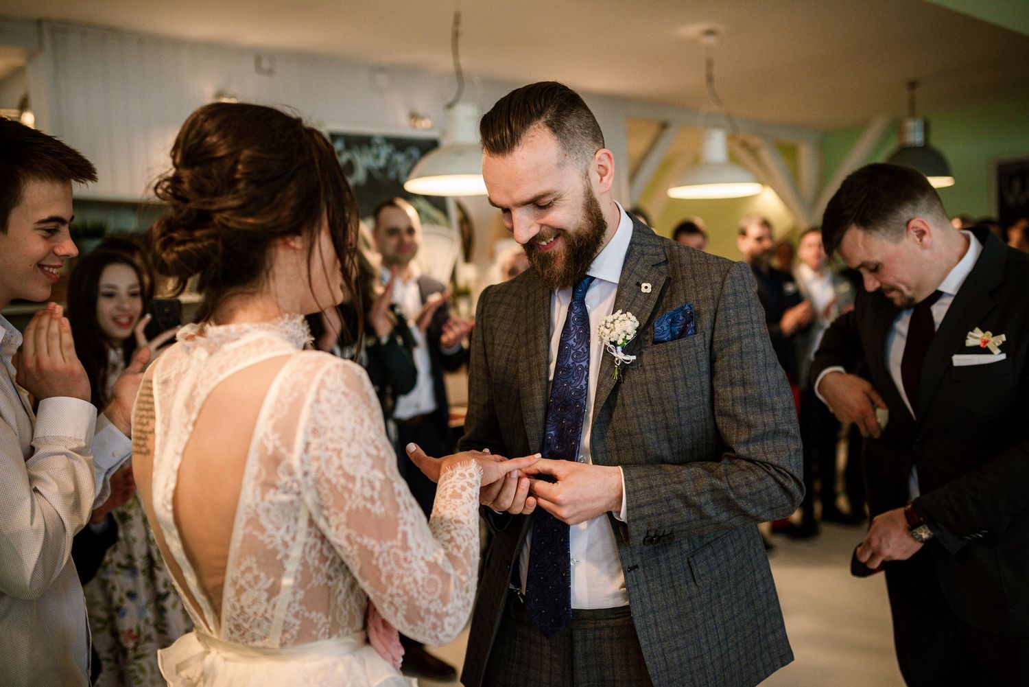Bearded groom in plaid suit places wedding ring on brides finger during ceremony.