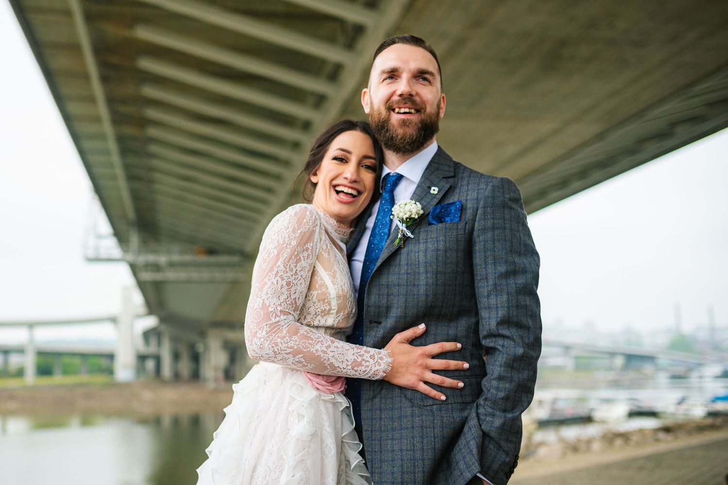 Joyful bride and groom laughing together under a bridge with a scenic waterway in the background.