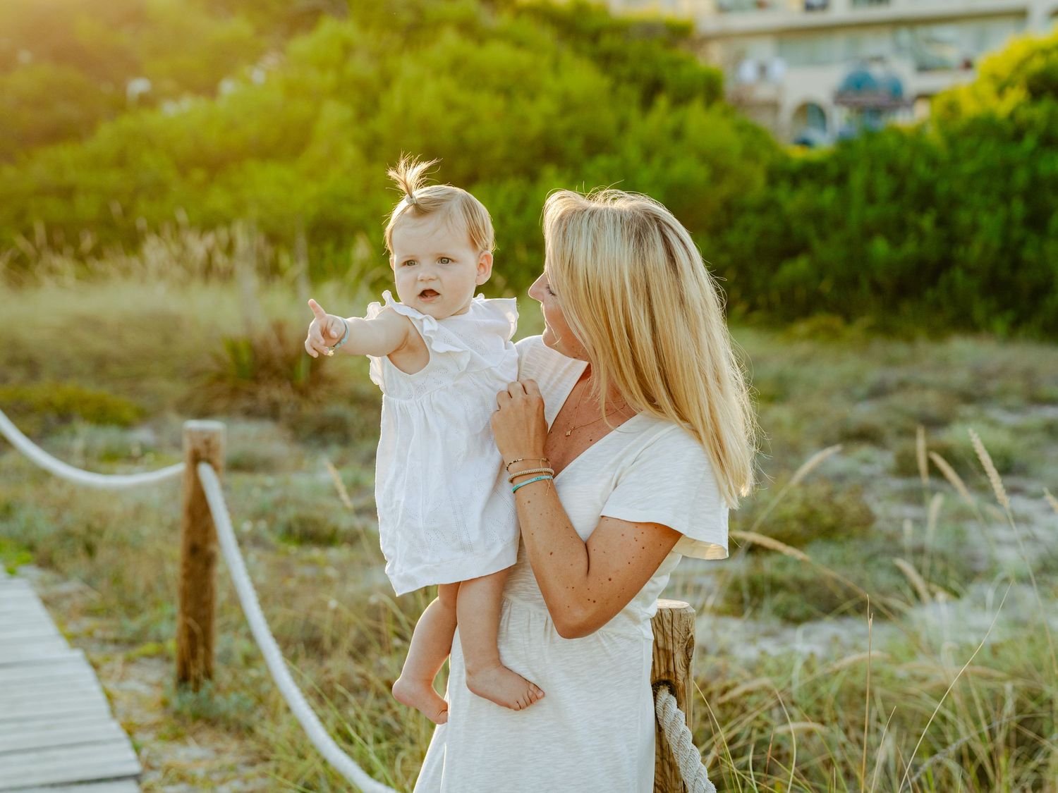 Blonde mother in white dress holding baby outdoors in golden light.