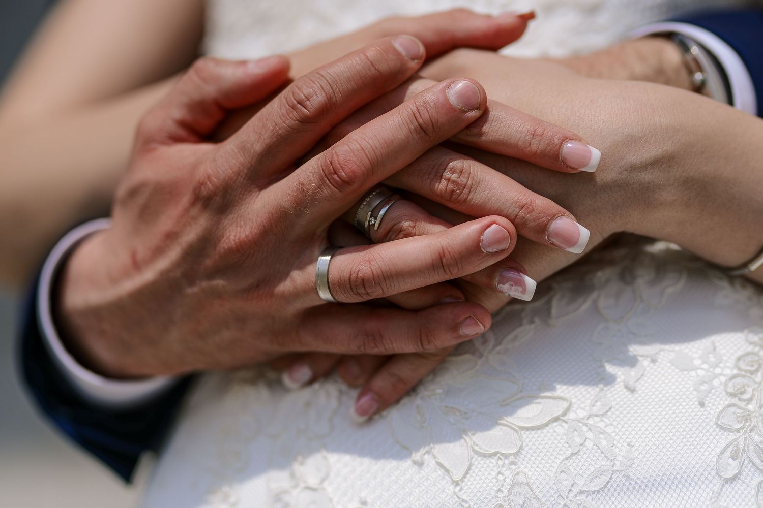 Close-up of bride and groom's hands with wedding rings resting on white lace wedding dress.