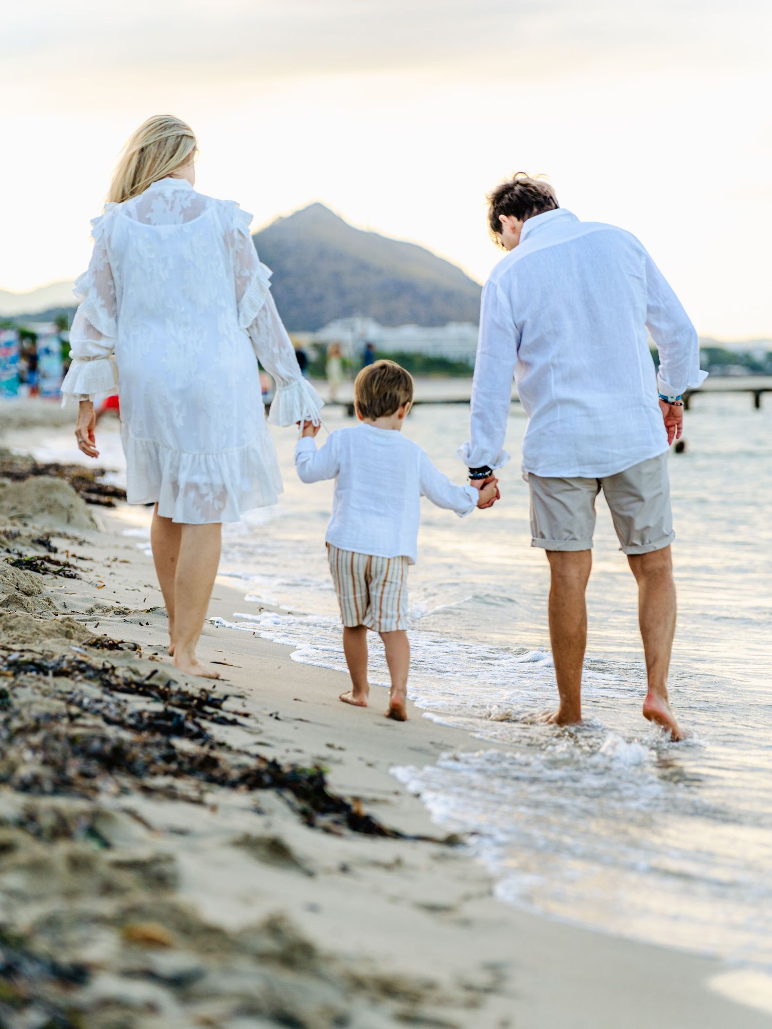 Family of three walking barefoot along a beach shoreline at sunset, holding hands from behind.