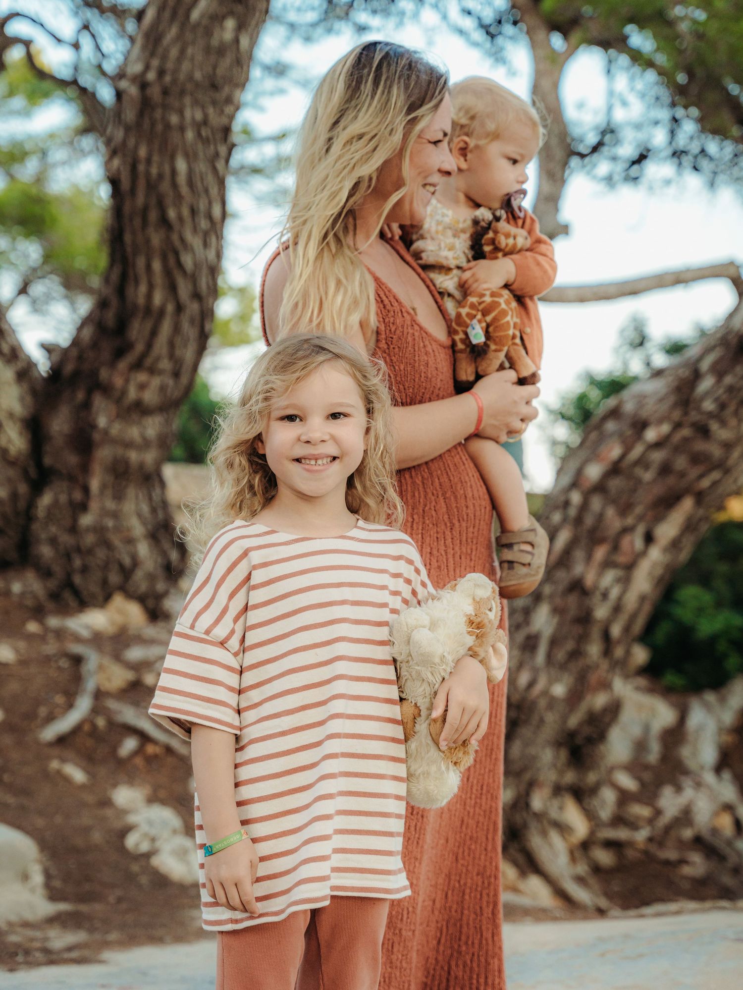 Blonde mother in rust dress holding baby near trees while young girl in striped shirt smiles at camera.