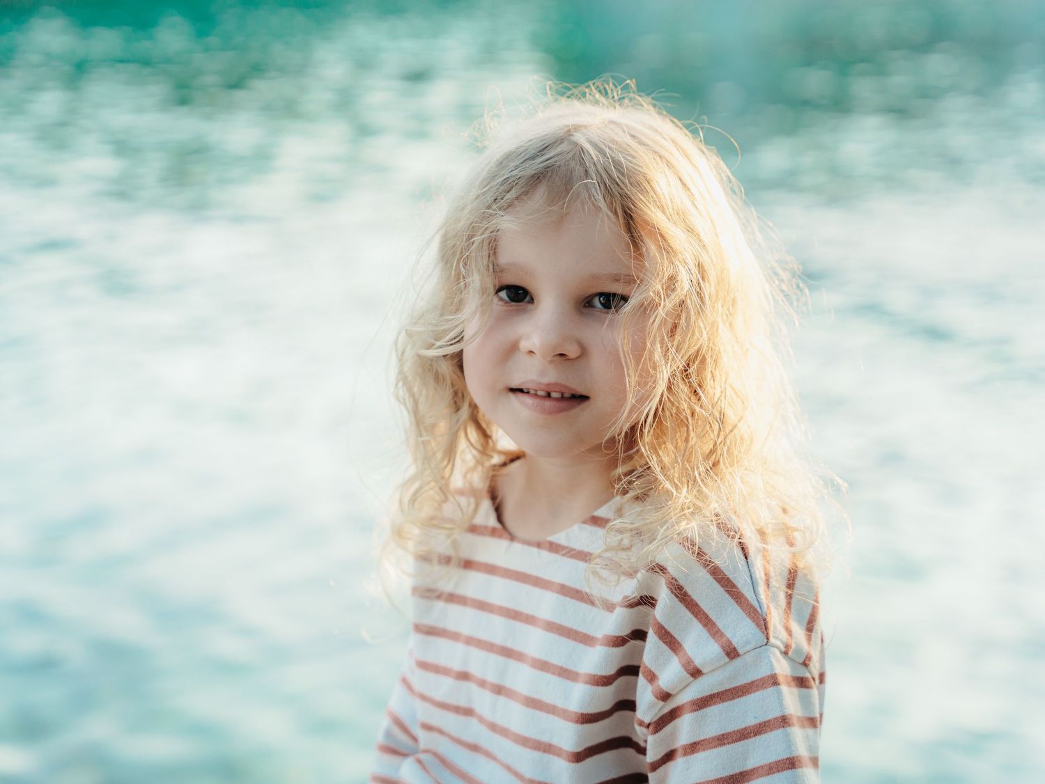 A young curly blonde girl in a striped shirt stands near shimmering turquoise water outdoors.