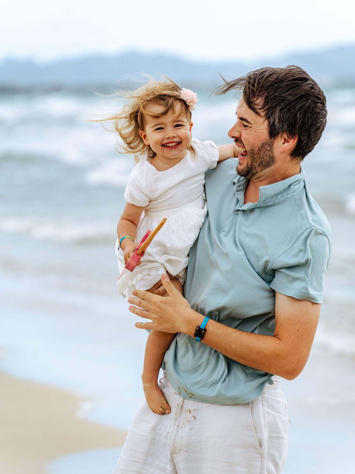 Happy father carrying his smiling toddler daughter with a pink flower on a beach.