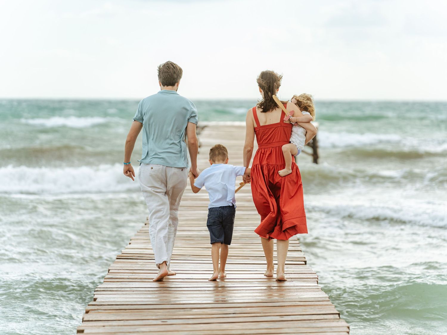 Family holding hands walking on wooden beach pier toward waves on a cloudy day.