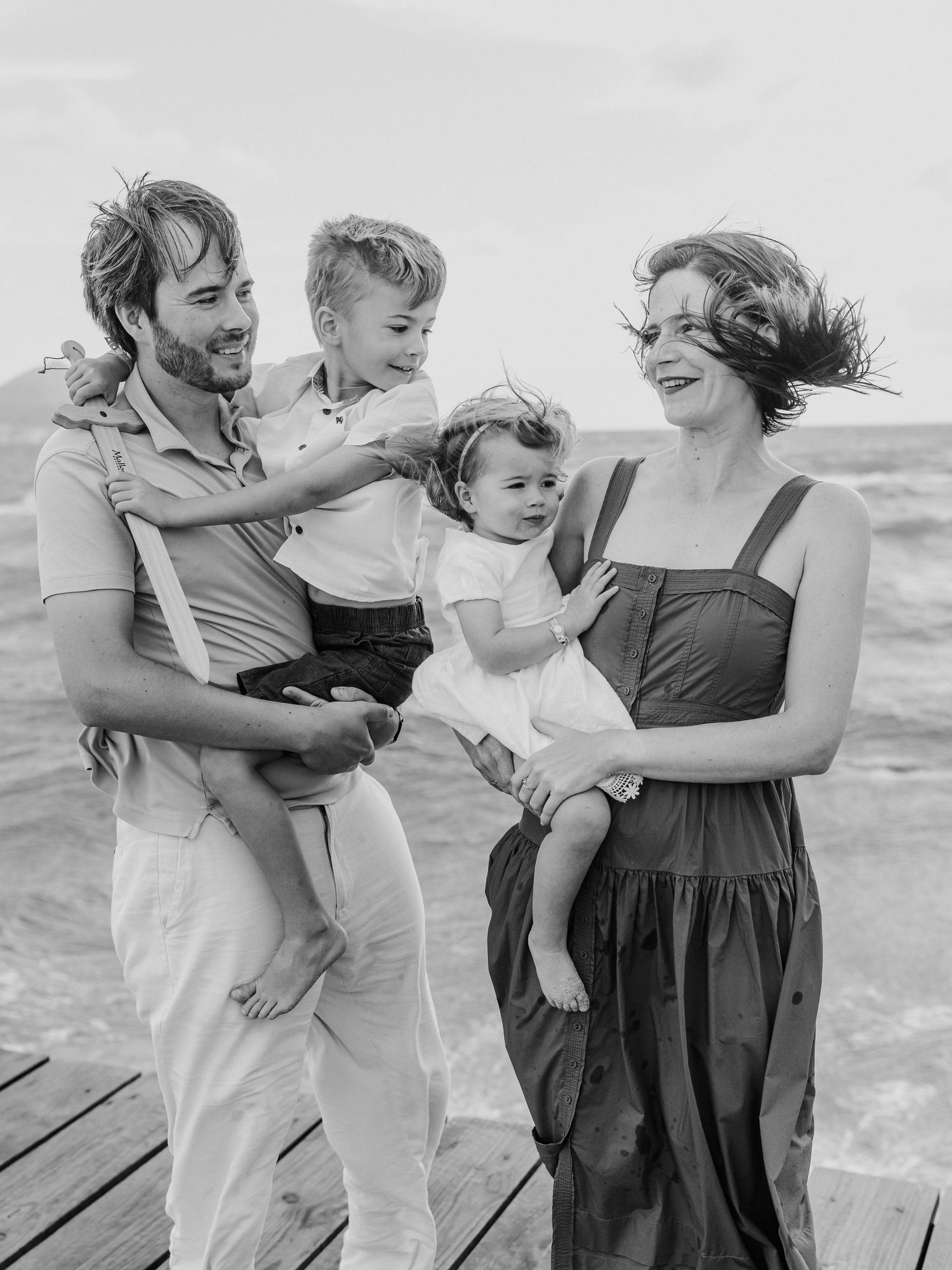 Black and white family photo on beach pier with parents holding two young children, wind blowing hair.
