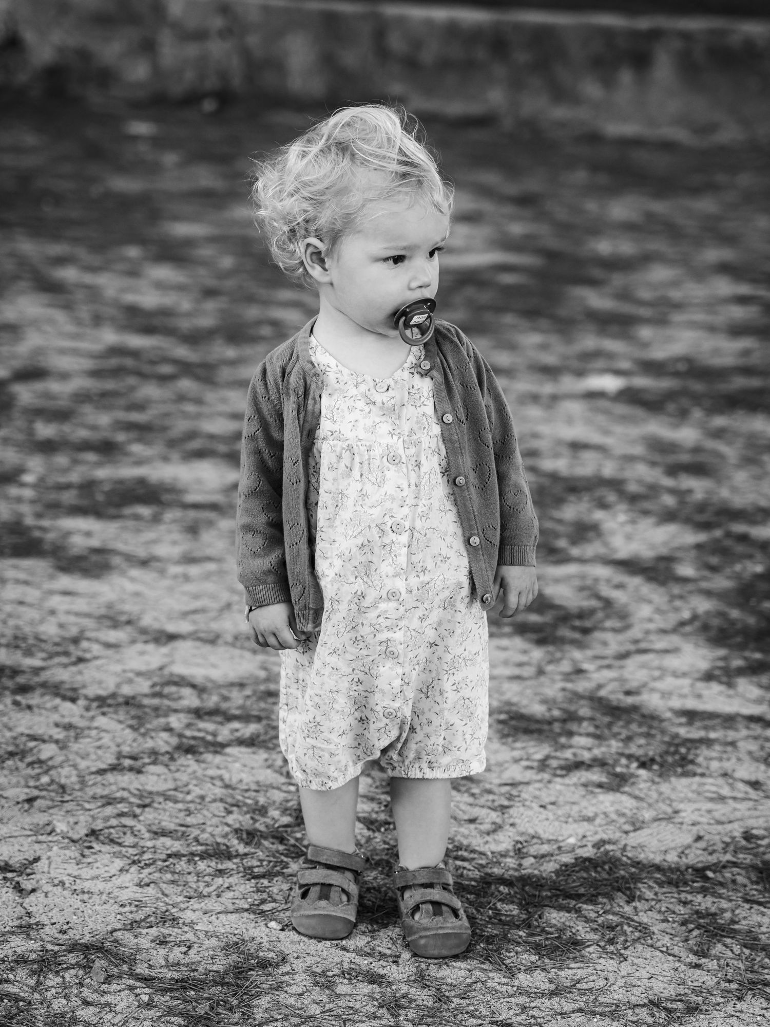 Black and white photo of toddler in floral dress and cardigan standing outdoors with pacifier.