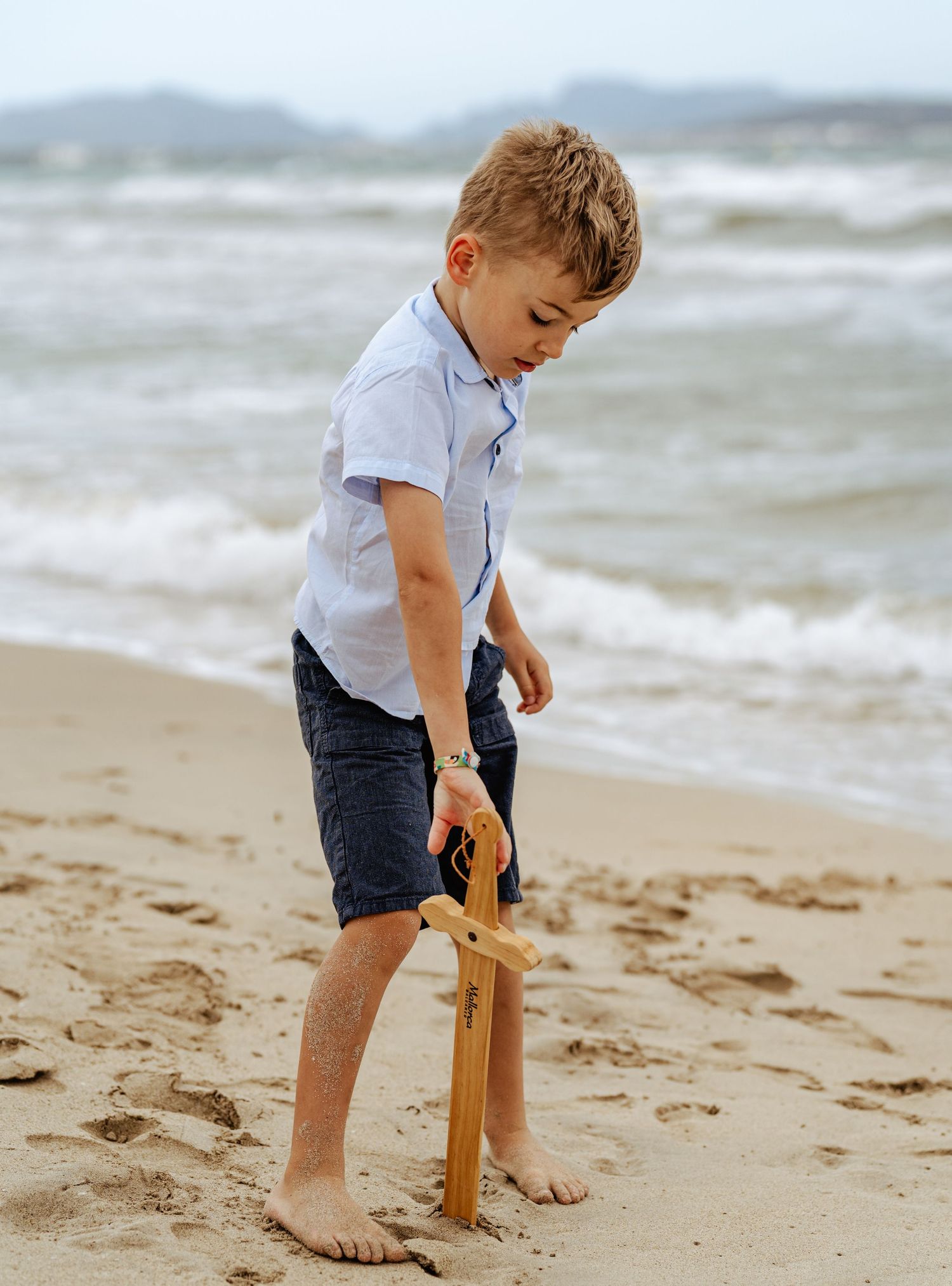 A young boy in a white shirt and shorts plays with a wooden toy sword on a sandy beach.